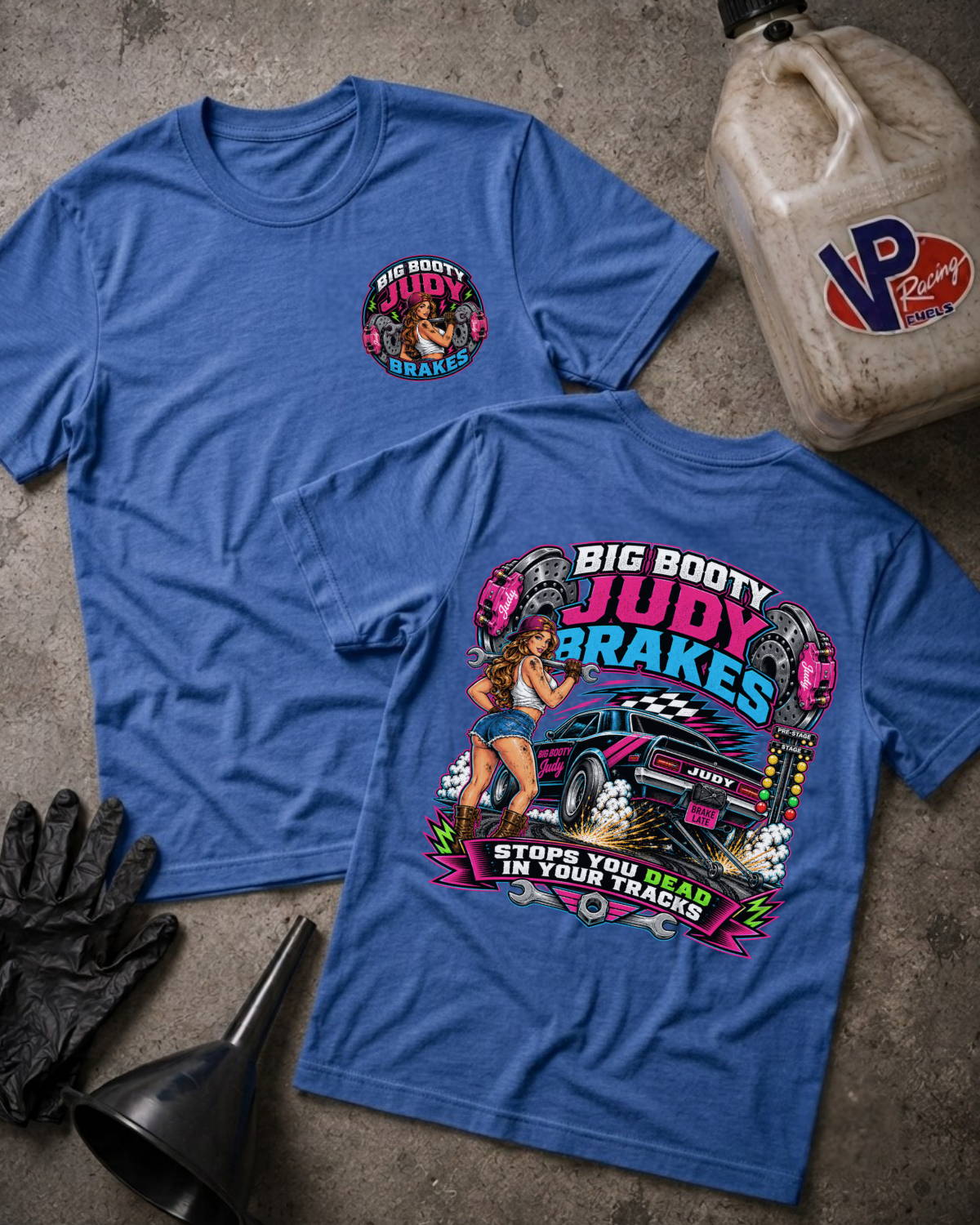 Two blue graphic T-shirts laid flat on a textured gray surface, with a black glove, funnel, and a VP Racing fuel can nearby. The front shirt shows a large colorful automotive pin-up graphic reading 'Big Booty Judy Brakes,' and the back shirt has a smaller matching circular chest graphic.