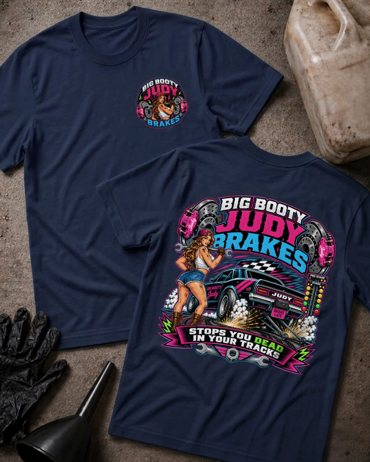 Two navy blue graphic t-shirts displayed flat on a textured gray surface, showing both the front and back designs. The front features a small circular chest graphic, while the back displays a large, colorful retro-style pin-up car graphic with the text 'BIG BOOTY JUDY BRAKES'.