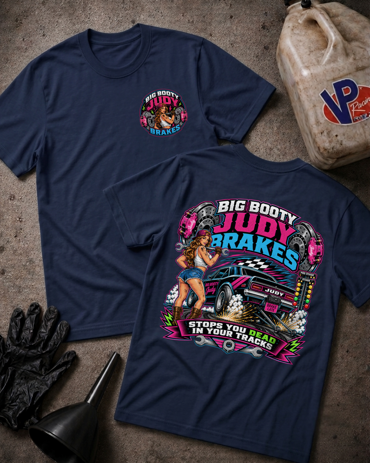 Two navy blue graphic T-shirts laid flat on a textured concrete surface, with a race gas jug, black gloves, and funnel as props. The front shirt shows a small multicolor chest graphic, and the back shirt shows a large colorful racing-themed print with a blue muscle car and the text 'BIG BOOTY JUDY BRAKES' and 'STOPS YOU DEAD IN YOUR TRACKS'.
