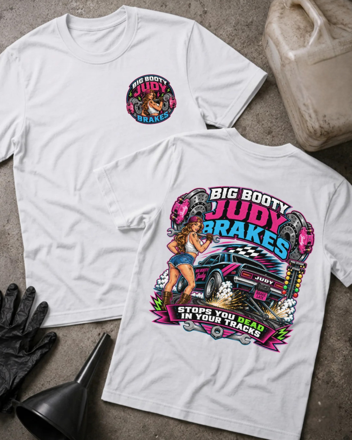 Two white graphic T-shirts laid flat on a concrete surface, with a black funnel, black gloves, and a plastic jug used as styling props. The front shirt shows a small circular chest graphic reading 'Big Booty Judy Brakes,' and the back shirt shows a large colorful racing-style graphic with a pin-up woman, a pink car, and bold text.