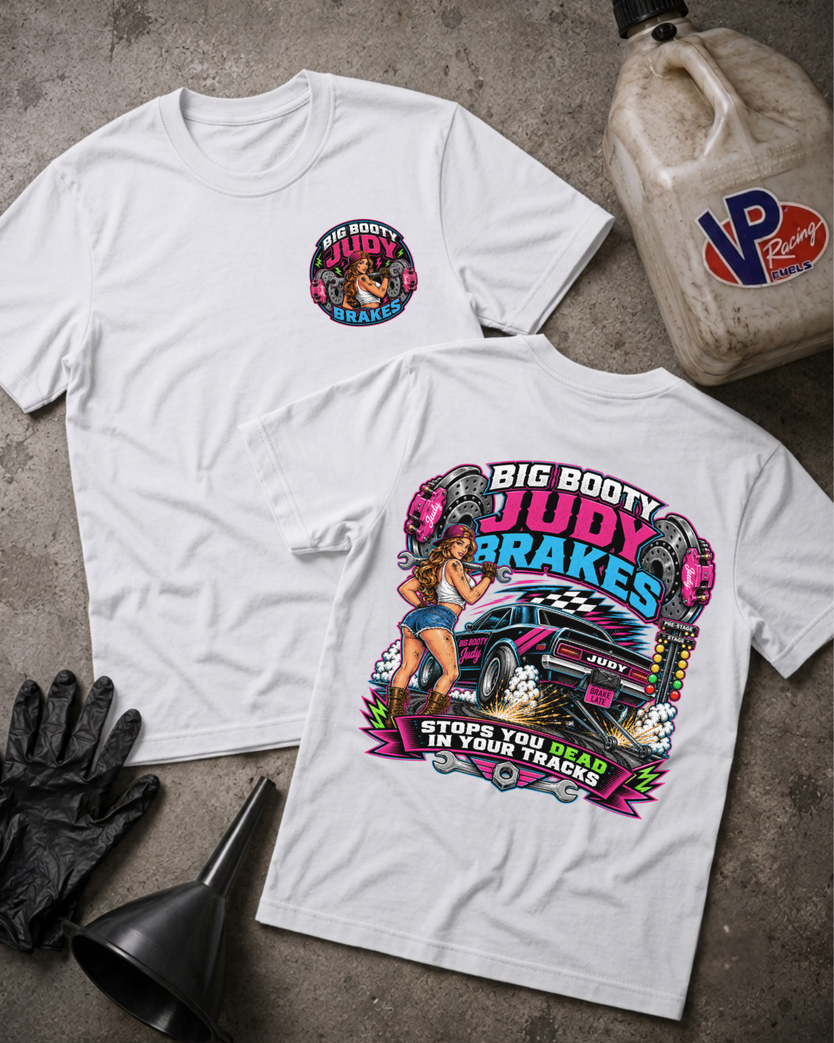 Two white graphic T-shirts laid flat on a gray concrete surface, with a black funnel, black gloves, and a VP Racing jug nearby. The front shirt has a small circular chest graphic, and the back shirt has a large colorful racing graphic with the text 'BIG BOOTY JUDY BRAKES' and 'STOPS YOU DEAD IN YOUR TRACKS'