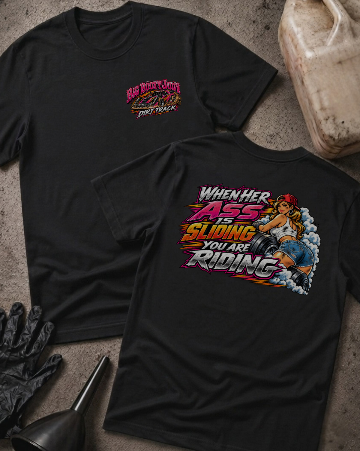 Big Booty Judy Dirt Track Racing T-shirt in black, flat lay showing the front chest logo and the back graphic reading When Her Ass Is Sliding You Are Riding, styled with work gloves, a funnel, and a fluid jug.