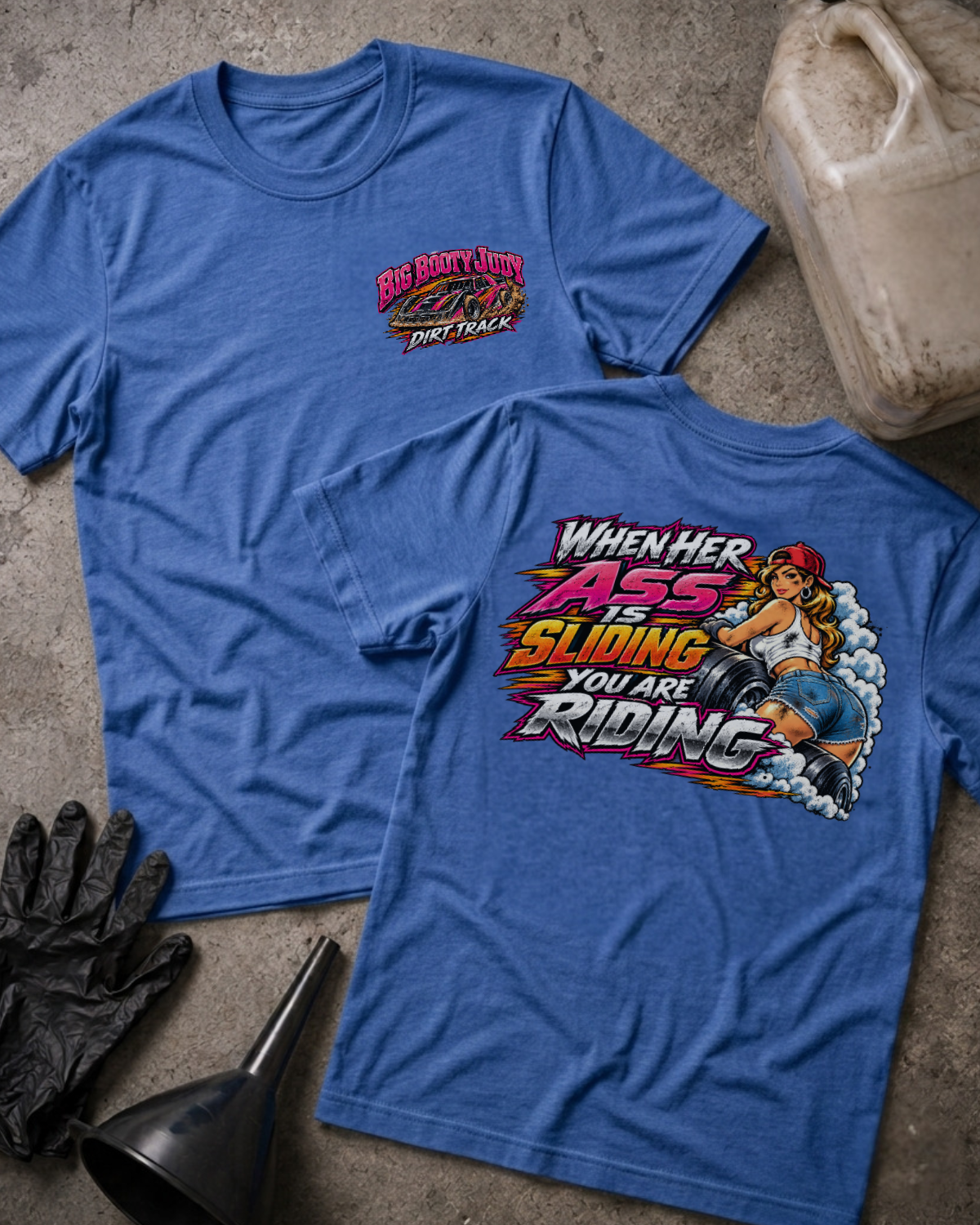 Big Booty Judy Dirt Track Racing T-shirt in heather true royal blue, flat lay showing the front chest logo and the back graphic reading When Her Ass Is Sliding You Are Riding, styled with work gloves, a funnel, and a fluid jug.