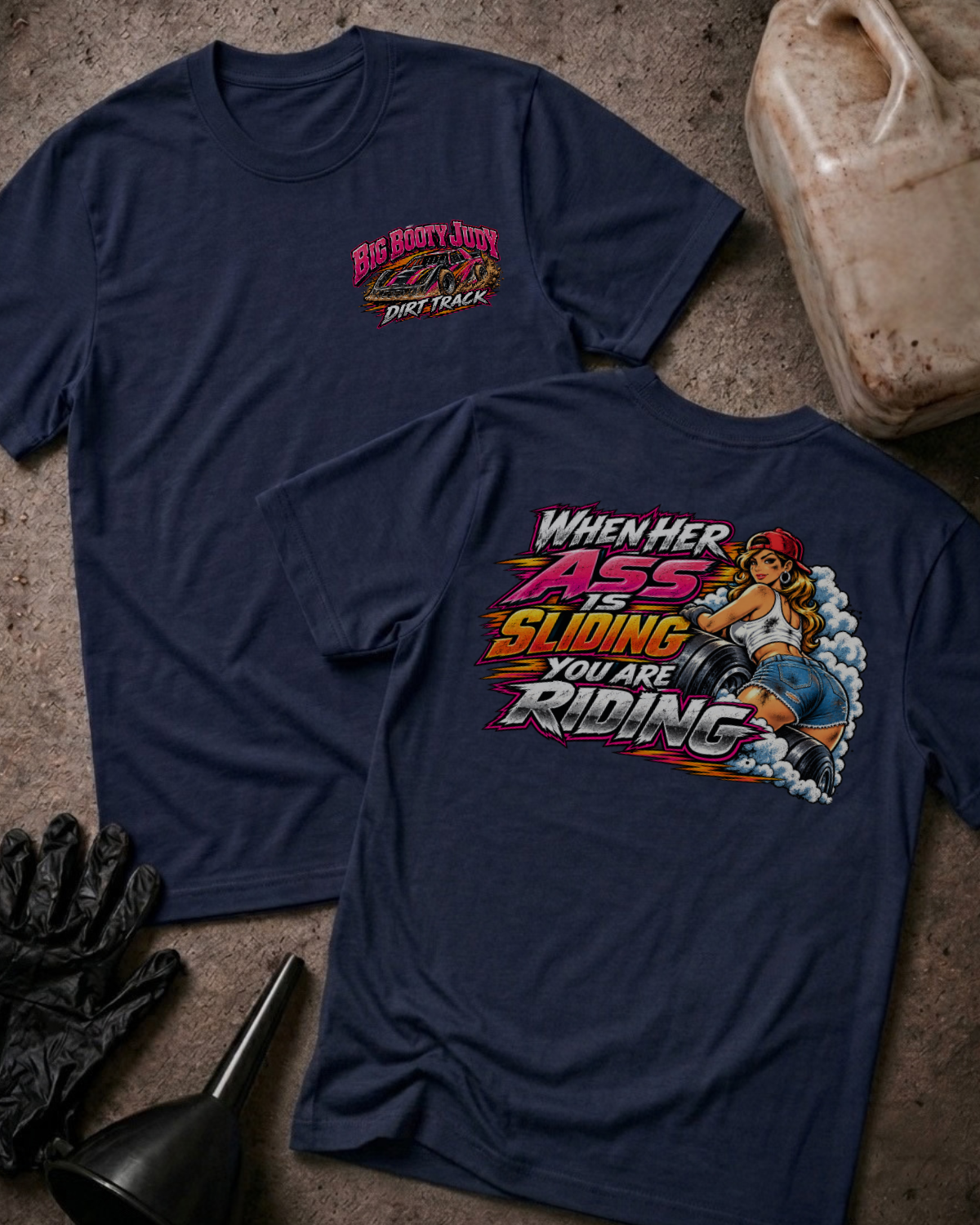 Big Booty Judy Dirt Track Racing T-shirt in navy, flat lay showing the front chest logo and the back graphic reading When Her Ass Is Sliding You Are Riding, styled with work gloves, a funnel, and a fluid jug.
