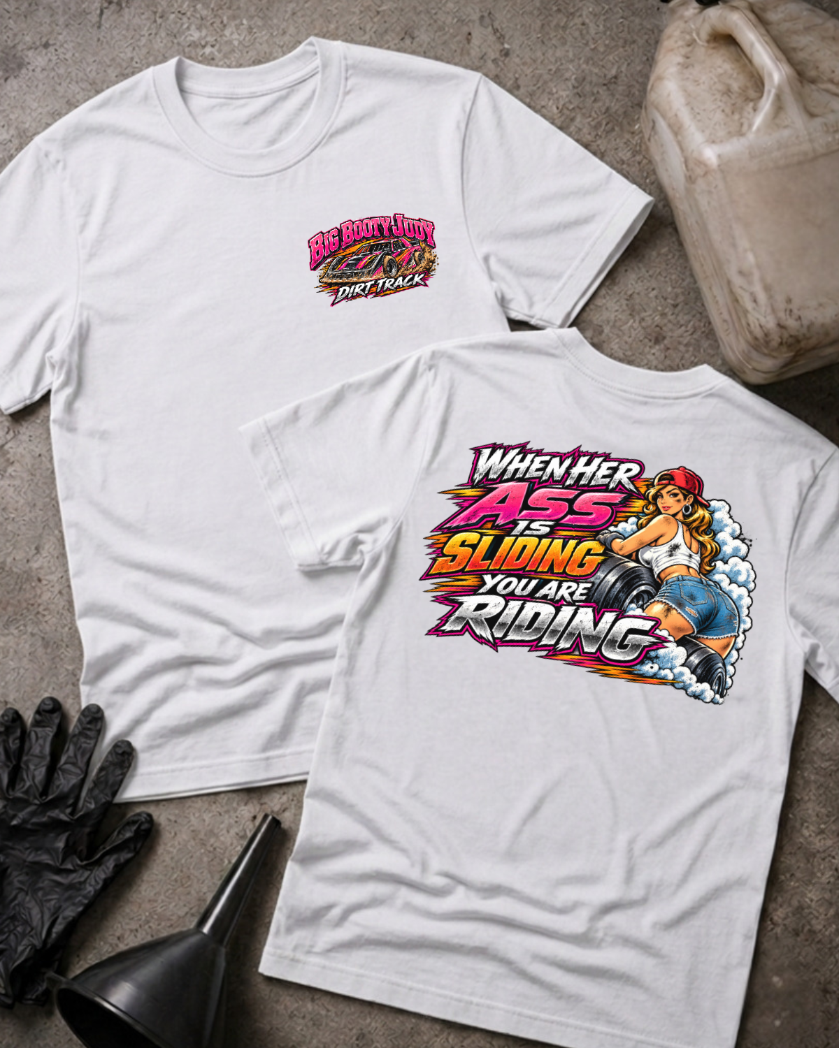 Big Booty Judy Dirt Track Racing T-shirt in white, flat lay showing the front chest logo and the back graphic reading When Her Ass Is Sliding You Are Riding, styled with work gloves, a funnel, and a fluid jug.