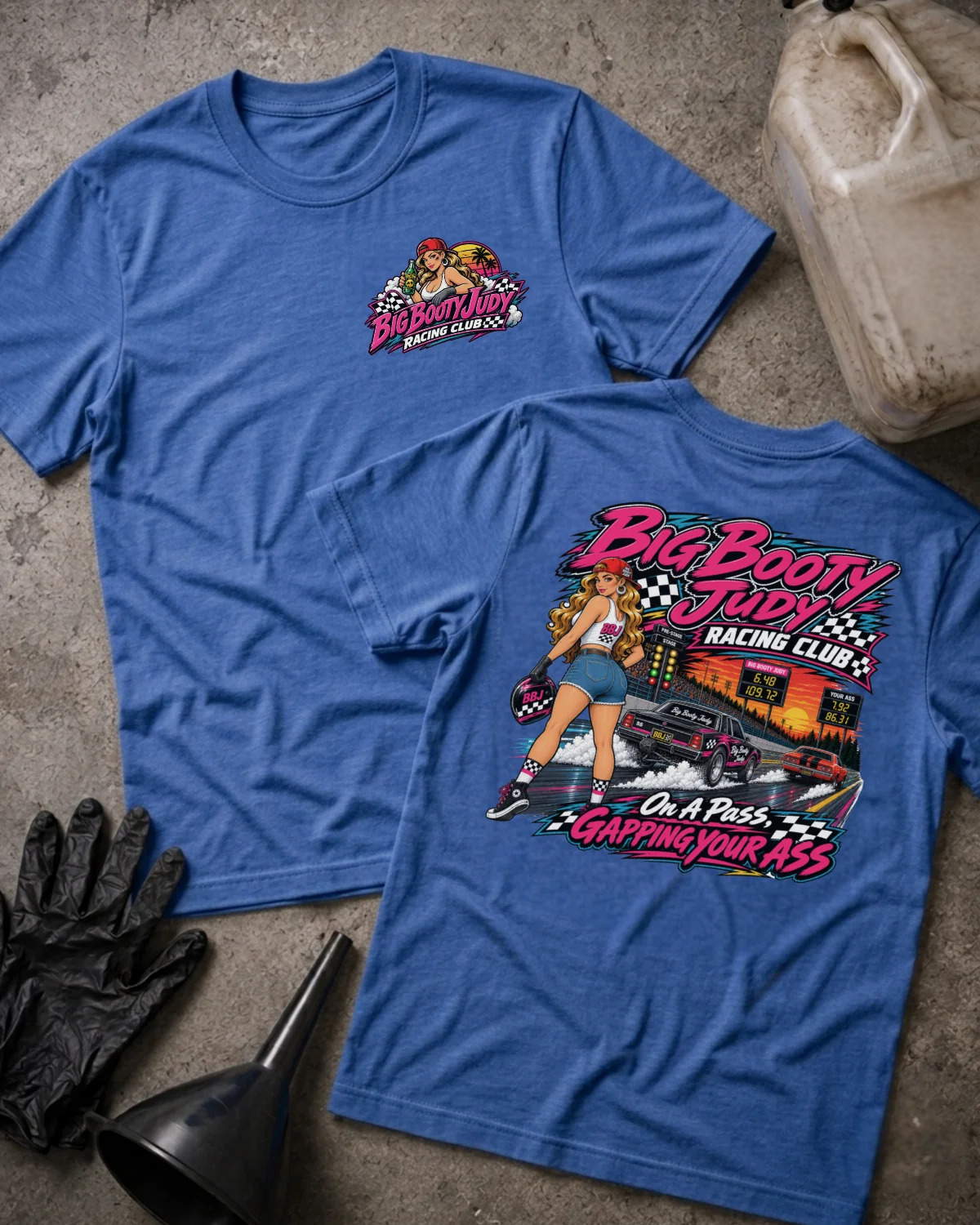 Two blue graphic T-shirts laid flat on a concrete surface, showing the front and back designs. The shirts feature a retro racing-themed print with a woman, race cars, checkered flags, and bold text reading 'Big Booty Judy Racing Club' and 'On A Pass Gapping Your Ass.