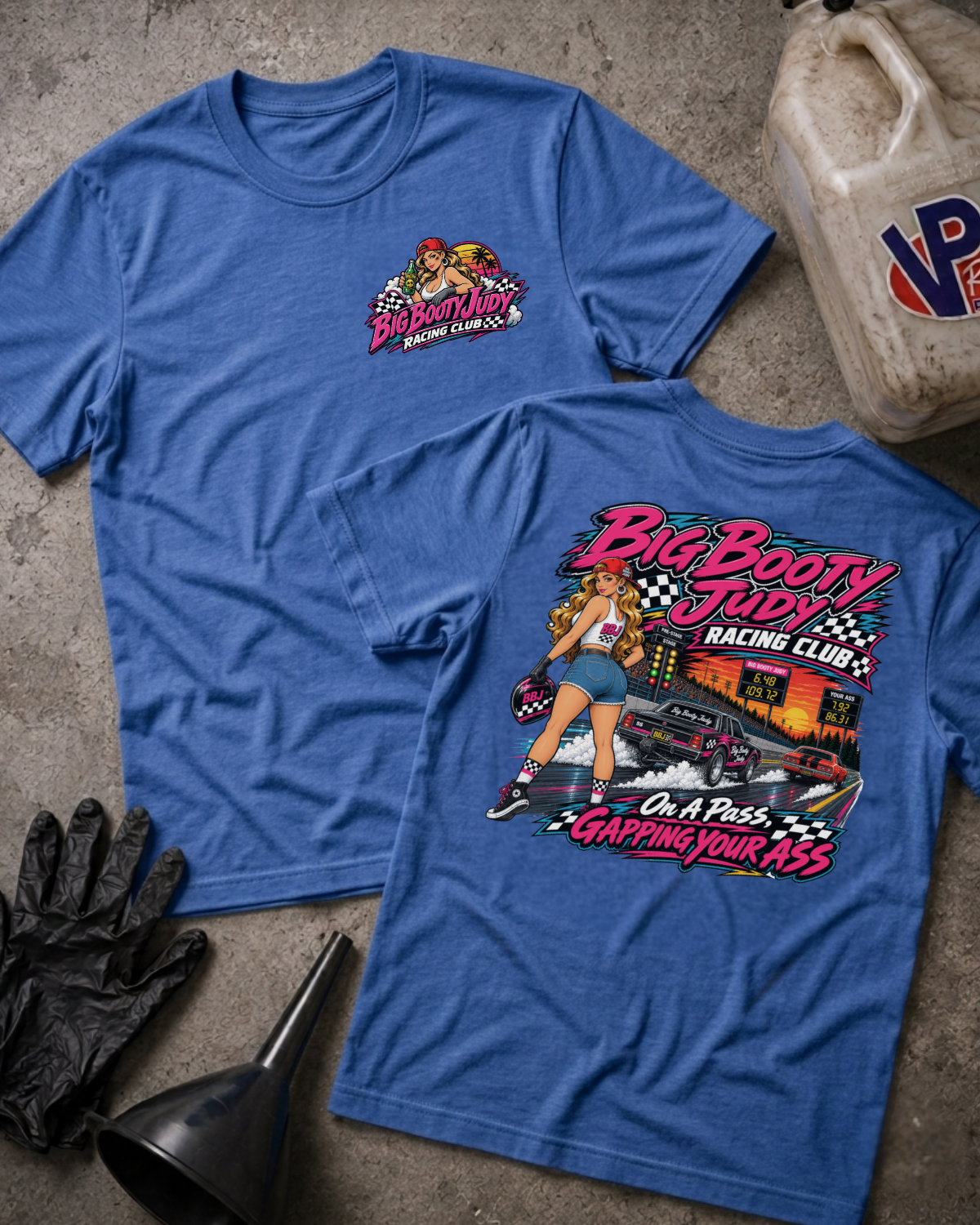 Two heather true royal graphic T-shirts displayed on a textured gray surface with racing-themed props, including a jerrycan, gloves, and funnel. The front shirt shows a small chest graphic, while the back shirt features a large colorful racing pin-up girl and race car graphic with the text 'Big Booty Judy Racing Club' and 'On A Pass Gapping Your Ass'.
