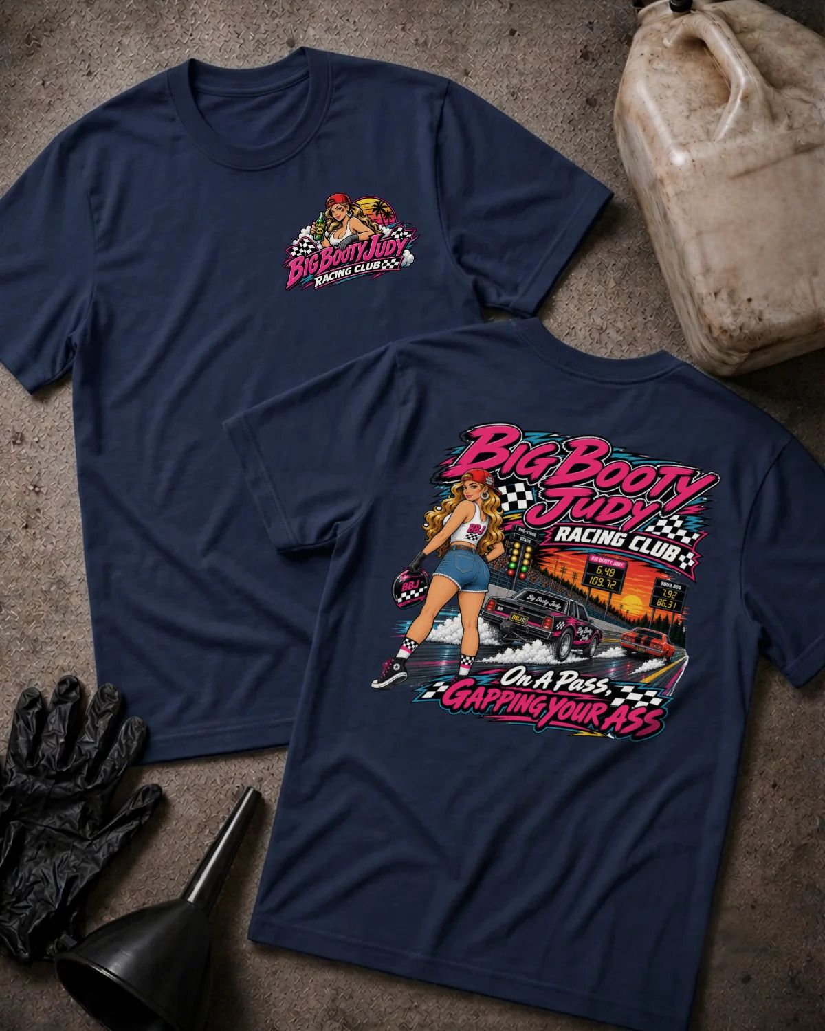 Two navy graphic T-shirts displayed on a textured gray surface with racing-themed props, including a jerrycan, gloves, and funnel. The front shirt shows a small chest graphic, while the back shirt features a large colorful racing pin-up girl and race car graphic with the text 'Big Booty Judy Racing Club' and 'On A Pass Gapping Your Ass'