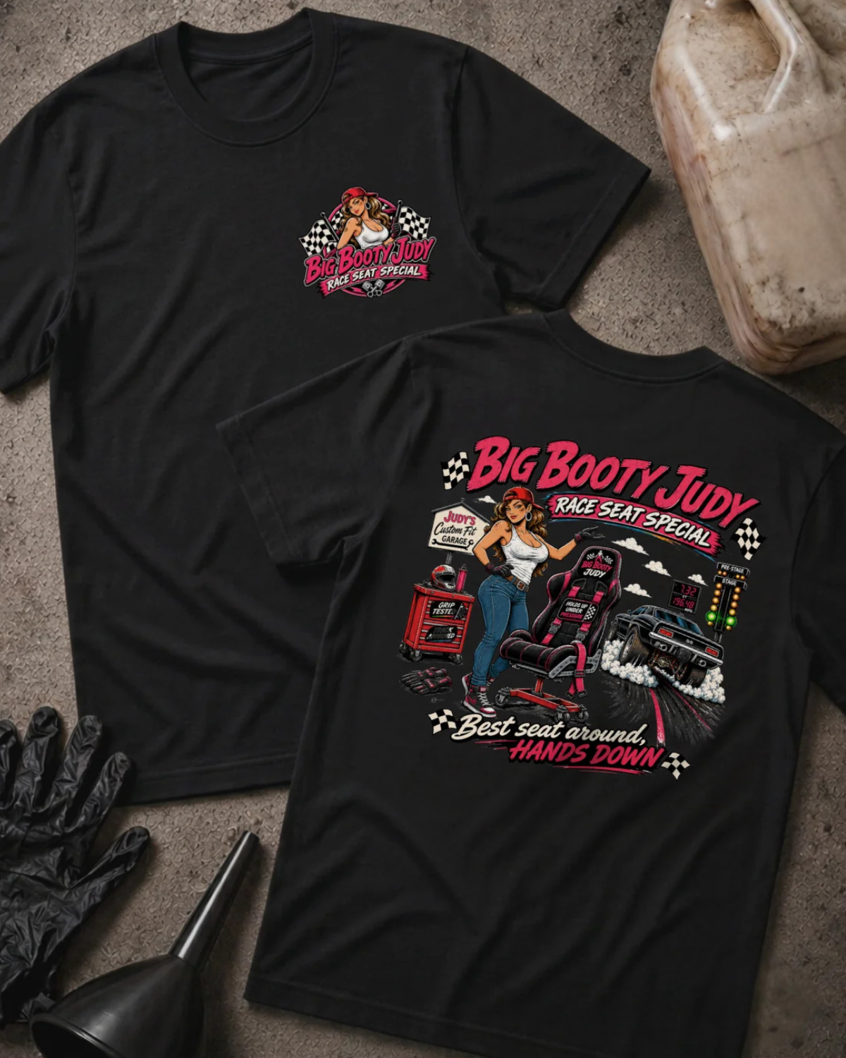 Two black graphic T-shirts displayed flat on a textured concrete surface, with a VP Racing jug, black gloves, and a black funnel as props. The front shirt shows a small red and white chest graphic, and the back shirt shows a large graphic of a woman in a racing seat with the text 'Big Booty Judy Take Seat Special' and 'Best seat around hands down'.