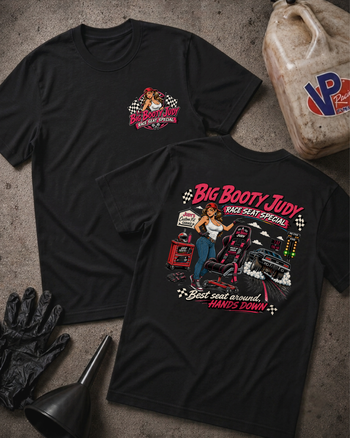 Two black graphic T-shirts laid flat on a gray concrete surface, with a large illustrated race car and the text 'Big Booty Judy Race Seat Special' on the back and a smaller matching chest graphic on the front. A plastic jug labeled VP Racing, black gloves, and a funnel are placed around them as styling props.