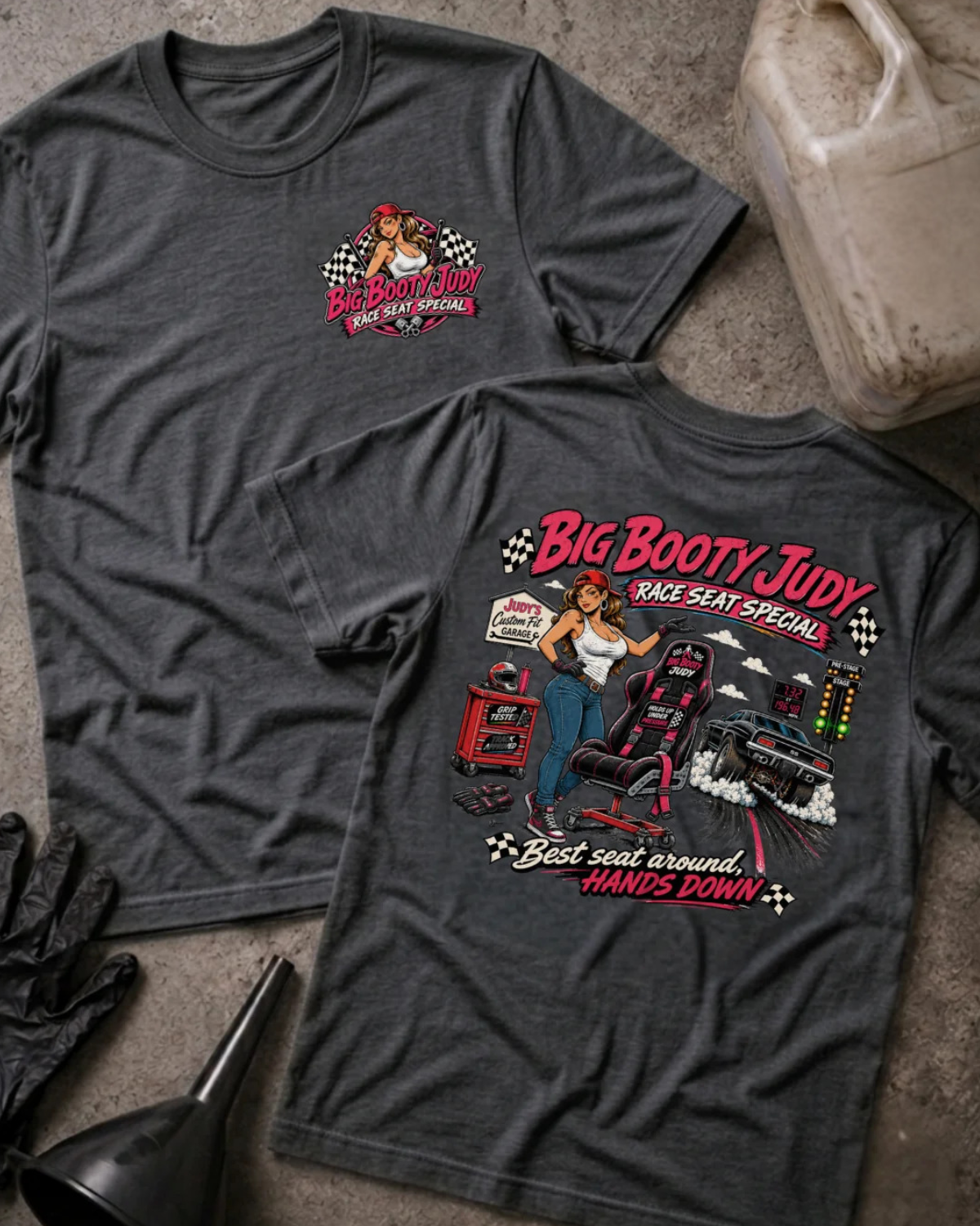 Two dark grey heather graphic T-shirts displayed flat on a textured concrete surface, with a VP Racing jug, black gloves, and a black funnel as props. The front shirt shows a small red and white chest graphic, and the back shirt shows a large graphic of a woman in a racing seat with the text 'Big Booty Judy Take Seat Special' and 'Best seat around hands down'.