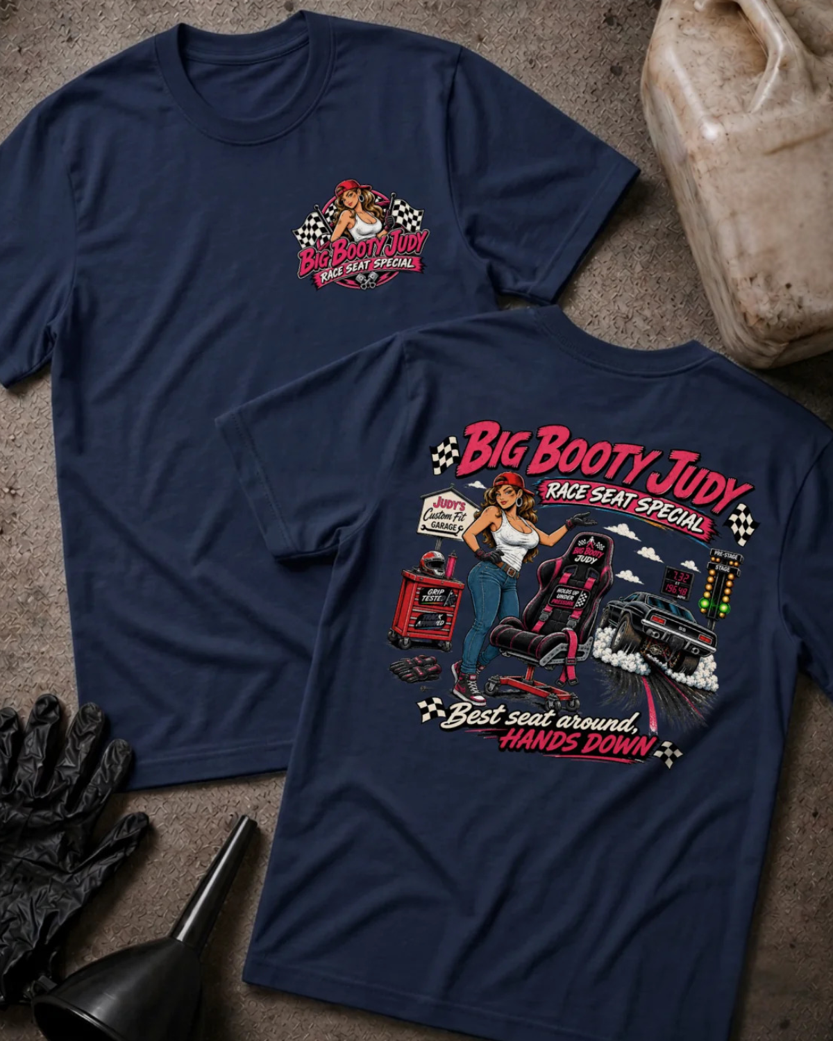 Two navy graphic T-shirts displayed flat on a textured concrete surface, with a VP Racing jug, black gloves, and a black funnel as props. The front shirt shows a small red and white chest graphic, and the back shirt shows a large graphic of a woman in a racing seat with the text 'Big Booty Judy Take Seat Special' and 'Best seat around hands down'.