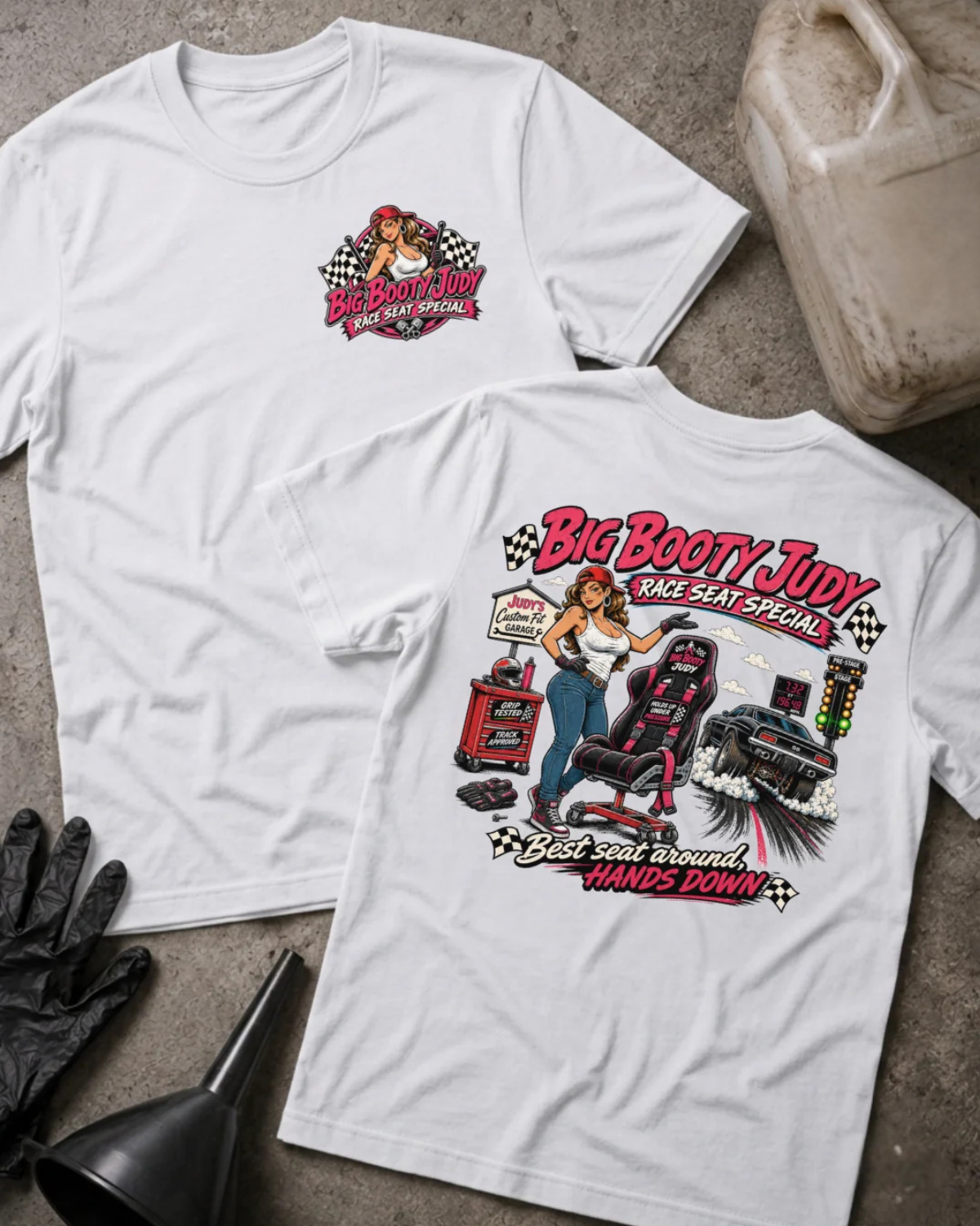 Two white graphic T-shirts displayed flat on a textured concrete surface, with a VP Racing jug, black gloves, and a black funnel as props. The front shirt shows a small red and white chest graphic, and the back shirt shows a large graphic of a woman in a racing seat with the text 'Big Booty Judy Take Seat Special' and 'Best seat around hands down'.
