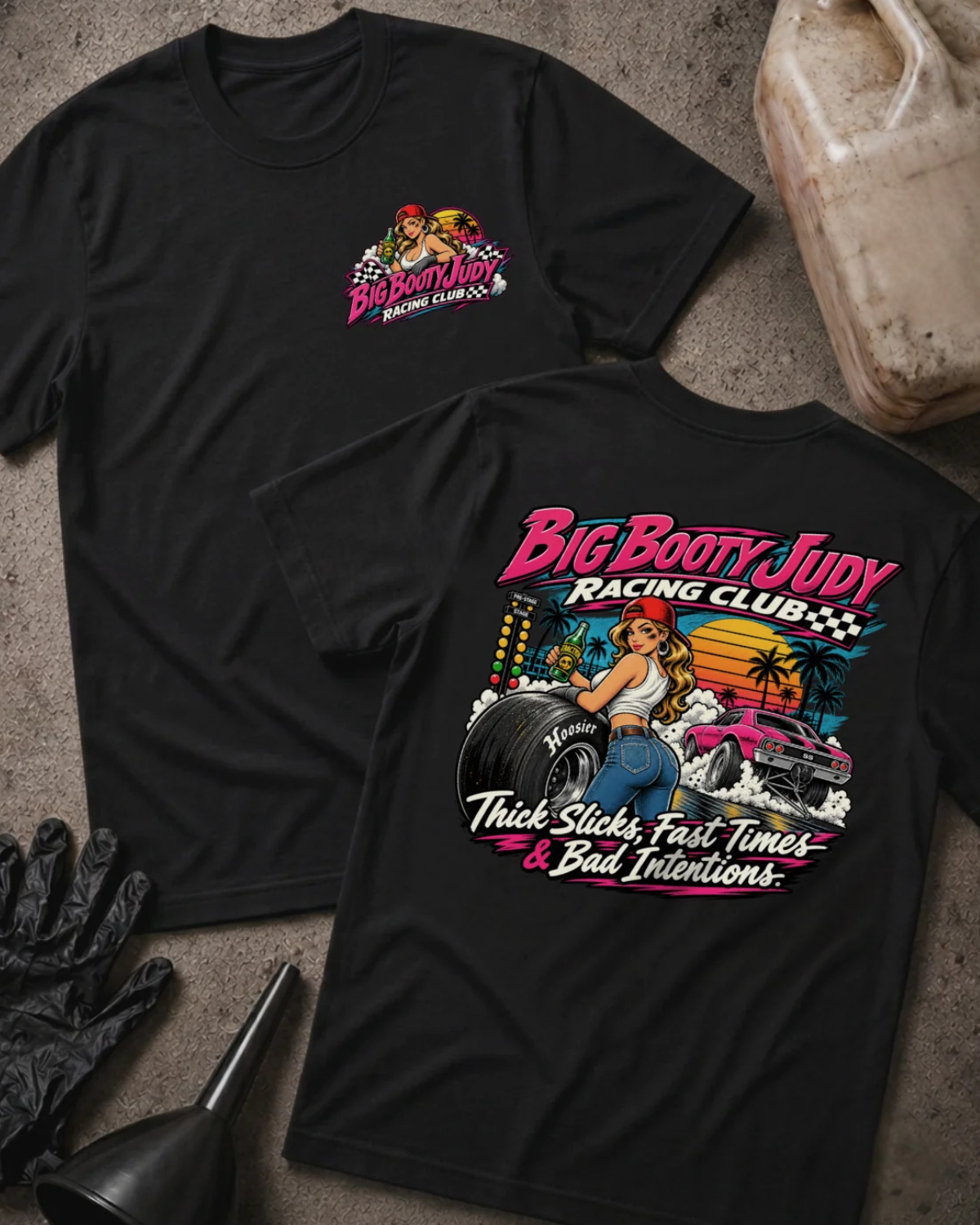 Two black graphic T-shirts laid flat on a textured gray surface, with racing-themed artwork and text. One shirt shows a small chest graphic, and the other shows a large back graphic with the words 'Big Booty Judy Racing Club' and 'Thick Slicks, Fast Time, and Bad Decisions'.