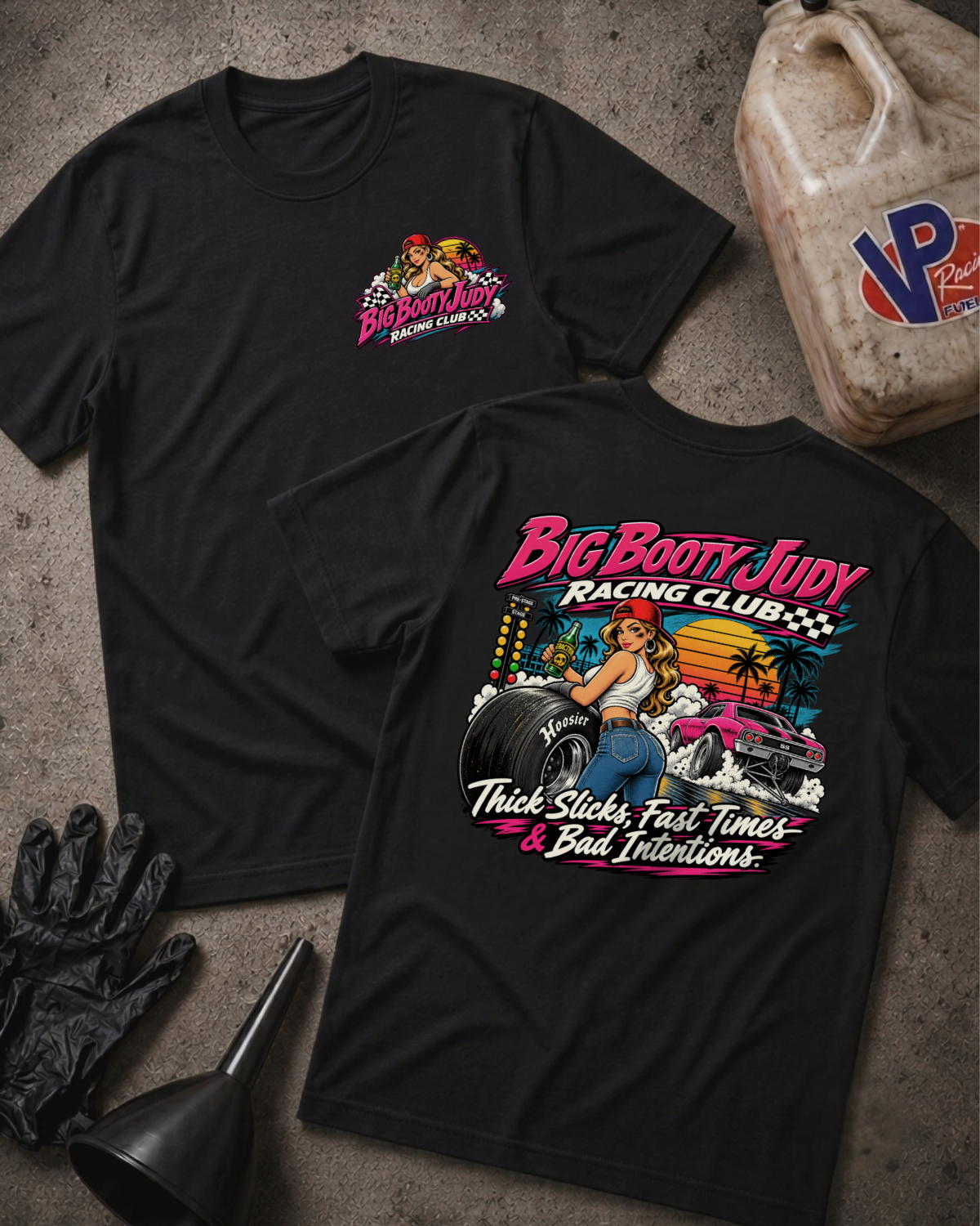 Two black graphic T-shirts laid flat on a textured gray surface, with racing-themed artwork and text. One shirt shows a small chest graphic, and the other shows a large back graphic with the words 'Big Booty Judy Racing Club' and 'Thick Slicks, Fast Time, and Bad Decisions'.