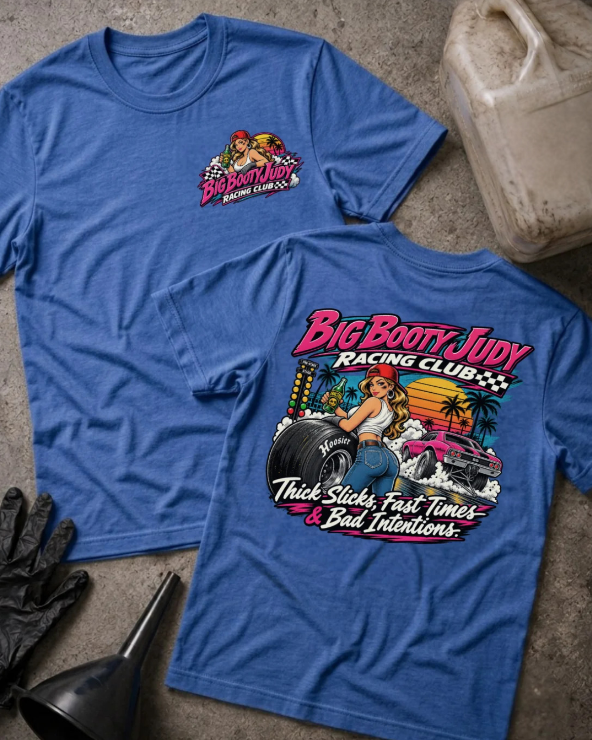 Two heather true royal graphic T-shirts laid flat on a textured gray surface, with racing-themed artwork and text. One shirt shows a small chest graphic, and the other shows a large back graphic with the words 'Big Booty Judy Racing Club' and 'Thick Slicks, Fast Time, and Bad Decisions'.