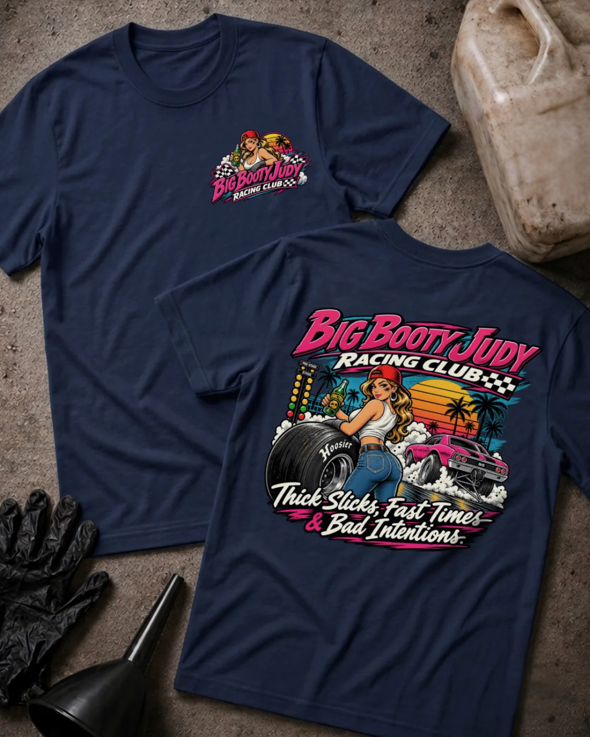 Two navy graphic T-shirts laid flat on a textured gray surface, with racing-themed artwork and text. One shirt shows a small chest graphic, and the other shows a large back graphic with the words 'Big Booty Judy Racing Club' and 'Thick Slicks, Fast Time, and Bad Decisions'.