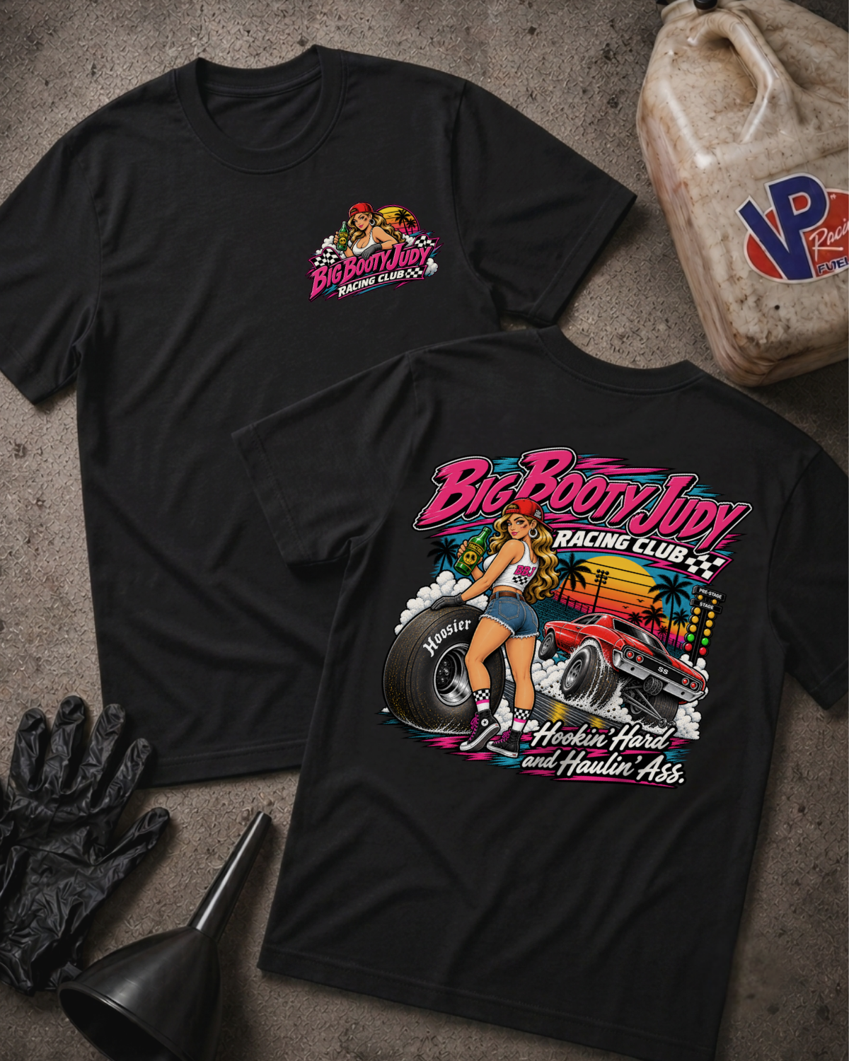 Two black graphic T-shirts laid flat on a textured gray surface, with racing-themed artwork and text. One shirt shows a small chest graphic, and the other shows a large back graphic with the words 'Big Booty Judy Racing Club' and 'Hookin' Hard and Haulin' Ass'