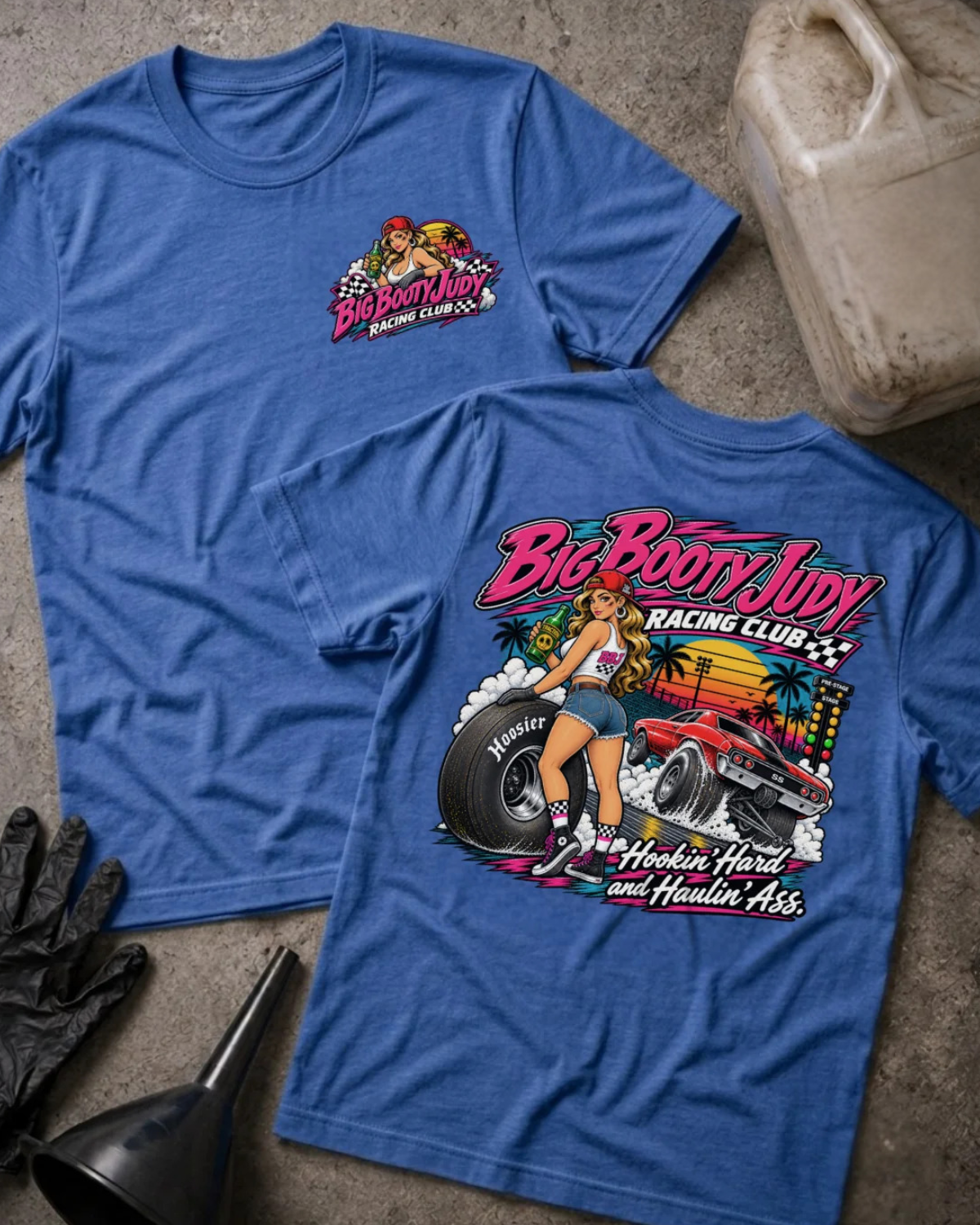Two heather true royal graphic T-shirts laid flat on a textured gray surface, with racing-themed artwork and text. One shirt shows a small chest graphic, and the other shows a large back graphic with the words 'Big Booty Judy Racing Club' and 'Hookin' Hard and Haulin' Ass'
