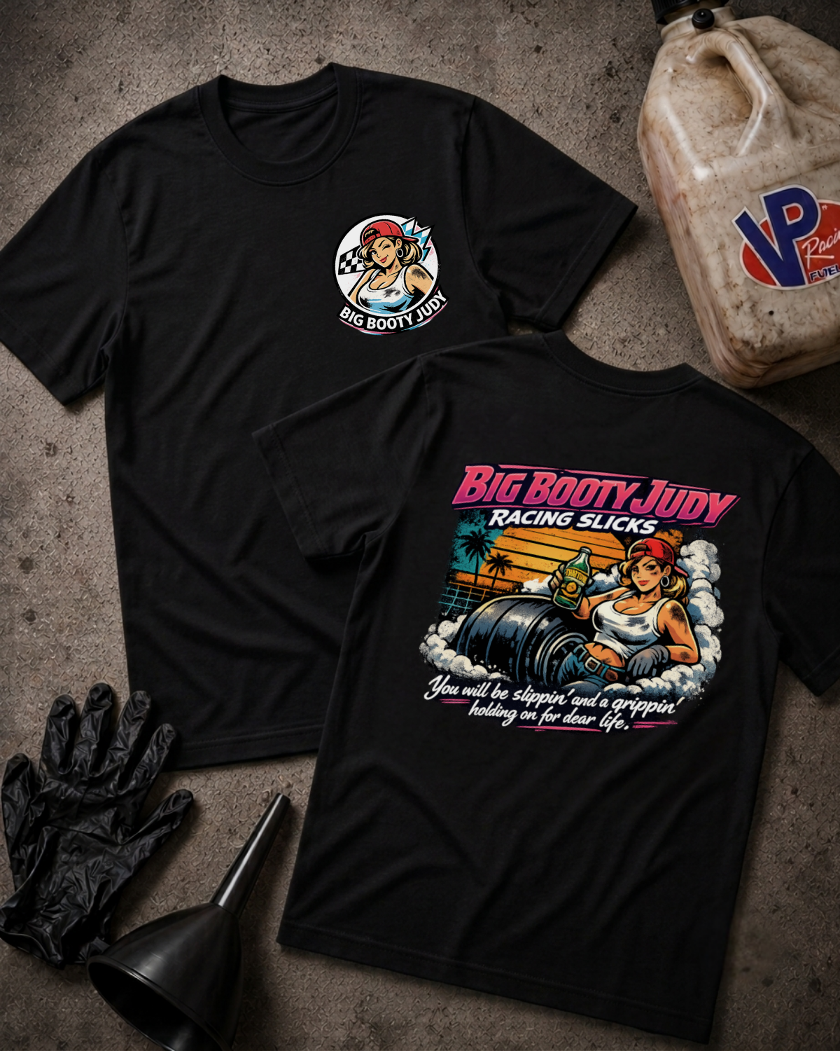 Two black graphic T-shirts laid flat on a textured gray surface, showing the front and back designs. The front has a small circular 'Big Booty Judy' chest graphic, and the back has a large colorful racing-themed print with the text 'Big Booty Judy Racing Slicks.