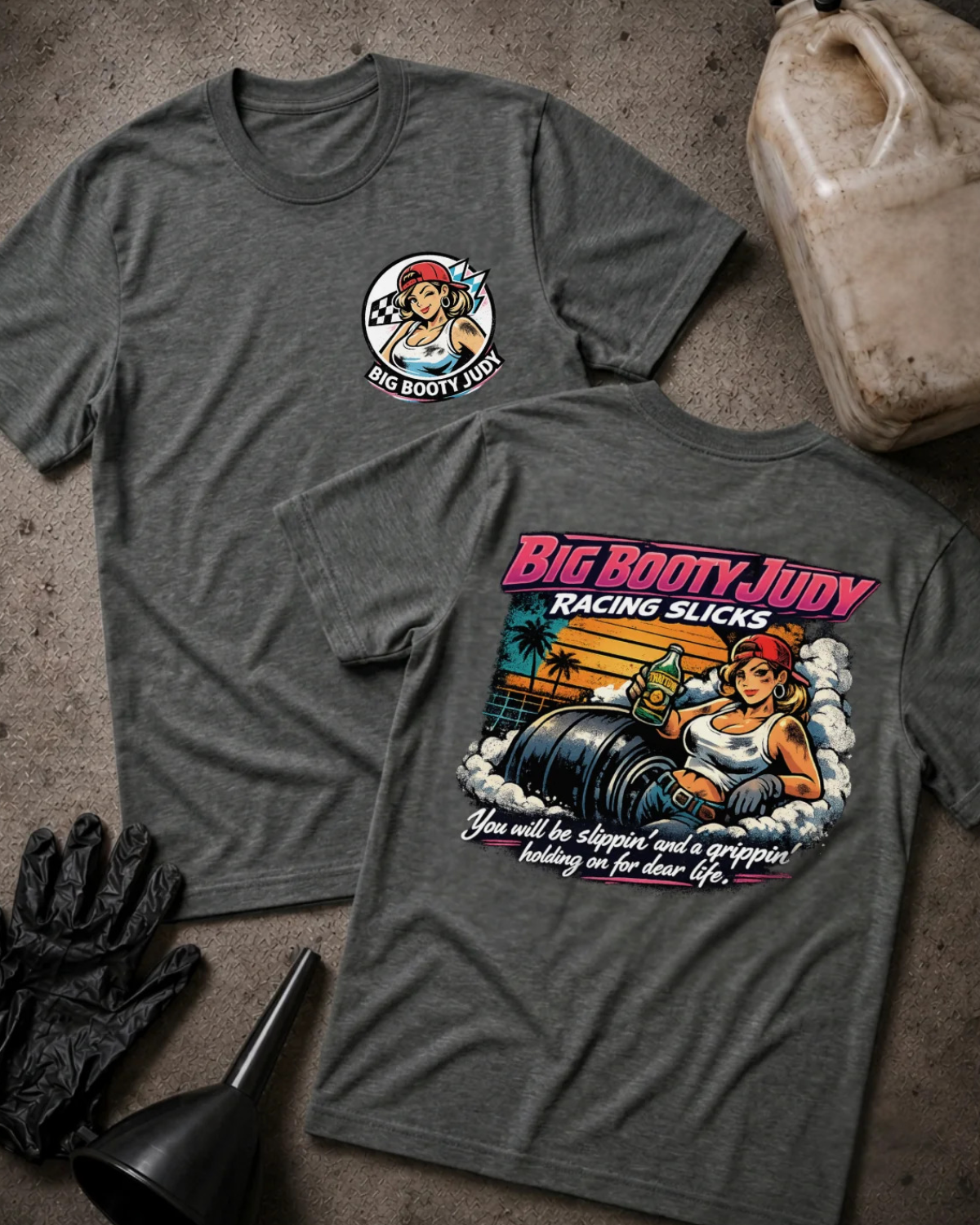 Two heather dark gray graphic T-shirts laid flat on a textured concrete surface, showing both front and back designs. The front has a small circular logo with a pin-up style illustration and the text 'BIG BOOTY JUDY'.