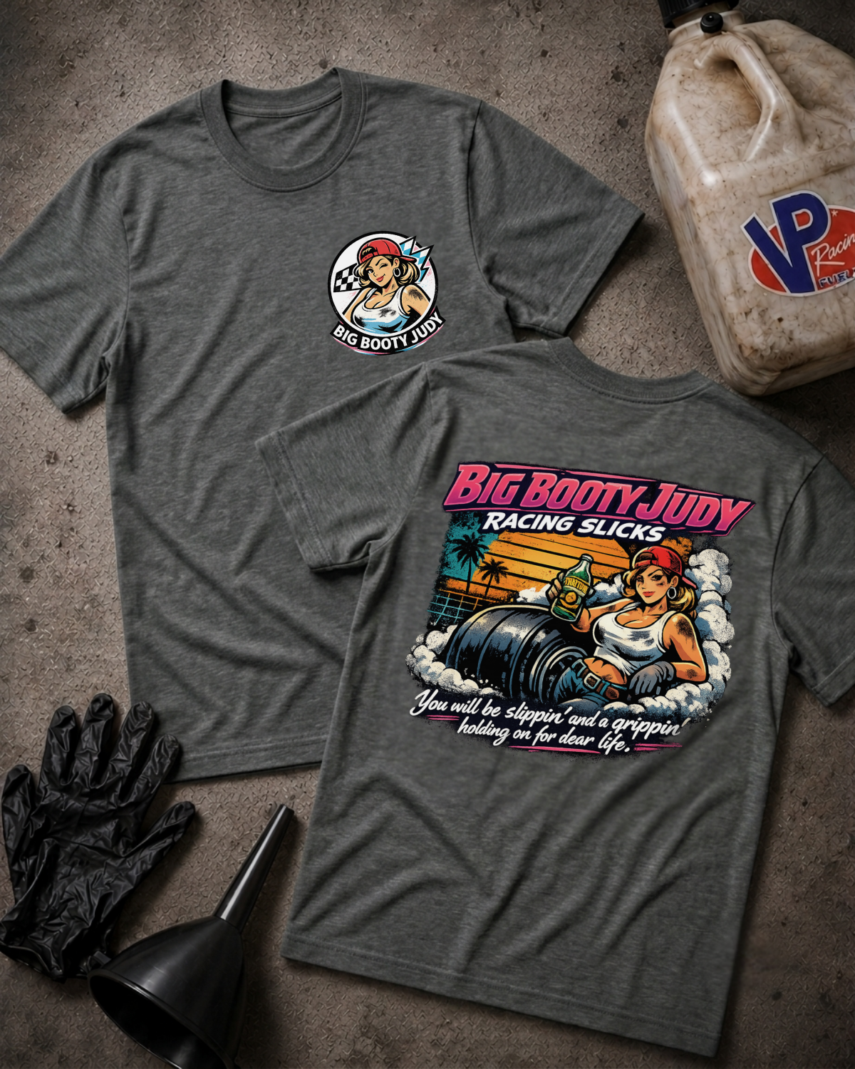 Two dark heather gray graphic T-shirts laid flat on a textured concrete-like surface, with black gloves and a dirty plastic jug as styling props. The front shirt shows a small circular pin-art graphic reading Big Rooty Judy, and the back shirt shows a large colorful racing-themed print with the same text and an illustrated woman beside racing sicks.