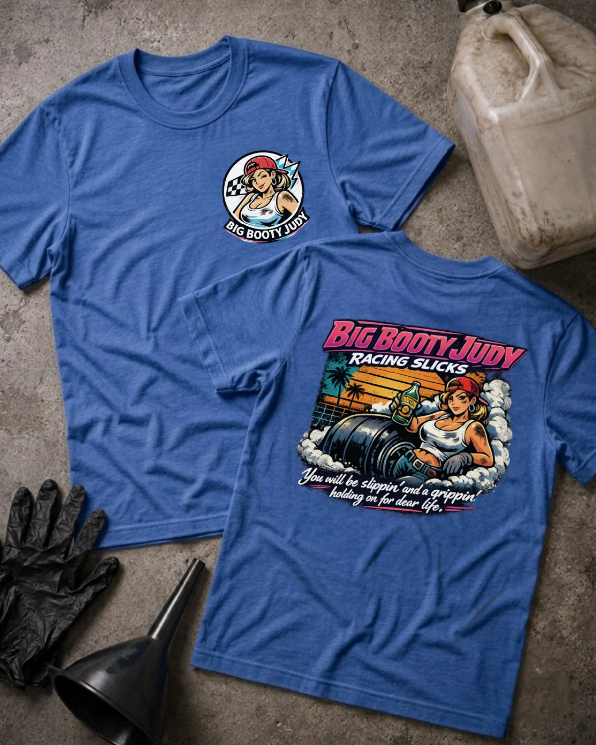 Two heather true royal black graphic T-shirts laid flat on a textured gray surface, showing the front and back designs. The front has a small circular 'Big Booty Judy' chest graphic, and the back has a large colorful racing-themed print with the text 'Big Booty Judy Racing Slicks.