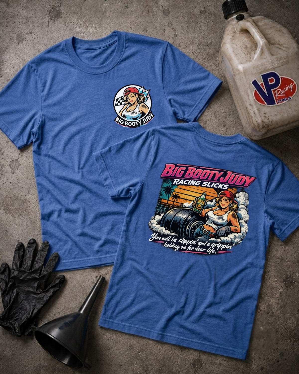 Two heather true royal graphic T-shirts laid flat on a textured gray surface, showing the front and back designs. The front has a small circular 'Big Booty Judy' chest graphic, and the back has a large colorful racing-themed print with the text 'Big Booty Judy Racing Slicks.
