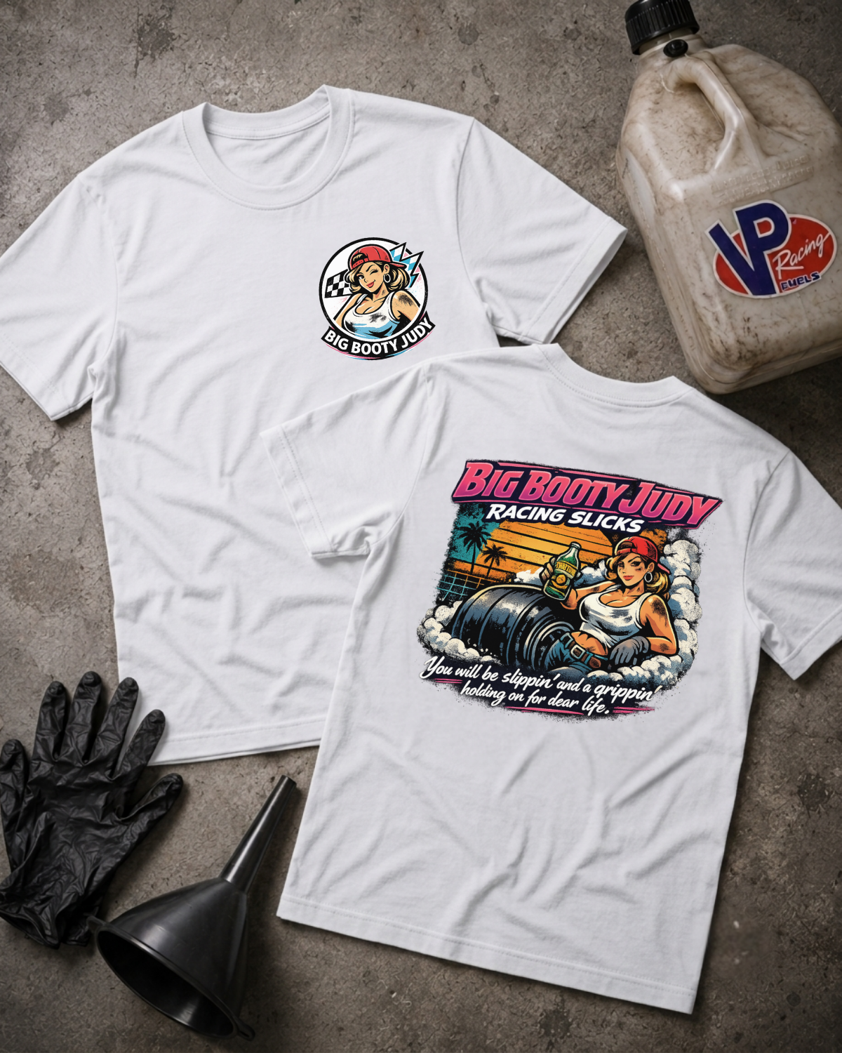 Two white graphic T-shirts laid flat on a textured gray surface, showing the front and back designs. The front has a small circular 'Big Booty Judy' chest graphic, and the back has a large colorful racing-themed print with the text 'Big Booty Judy Racing Slicks.