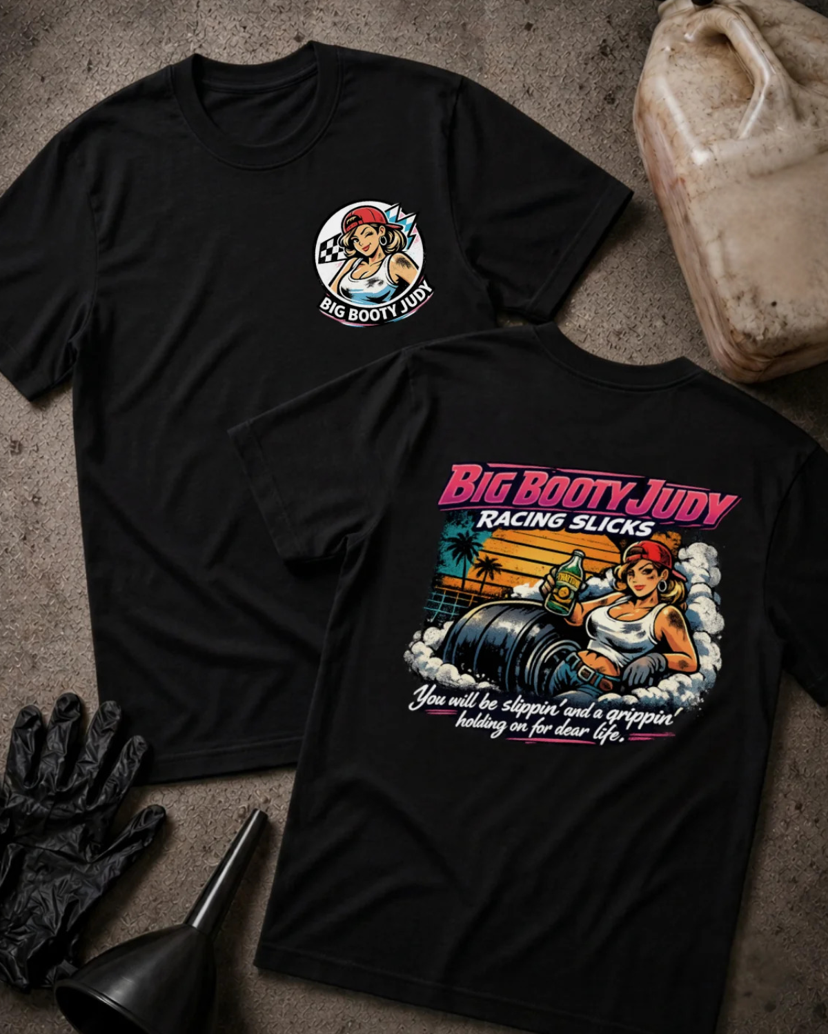 Two black graphic T-shirts laid flat on a textured gray surface, with racing-themed artwork and the text 'BIG BOOTY JUDY RACING SLICKS.' One shirt shows a small circular chest graphic, and the other shows a large back graphic with a pin-up style illustration and colorful text.