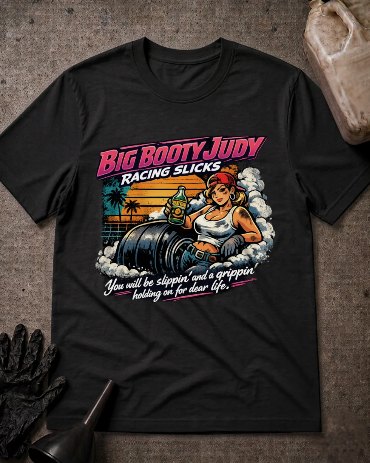 A black  graphic t-shirt laid flat on a textured gray surface, surrounded by racing-themed props including a black glove, funnel, and a jug of VP Racing. The shirt features a retro-style graphic of a woman in a racing suit with the text 'BIG BOOTY JUDY RACING SLICKS' and 'You will be slippin' and a grippin holding on for dear life.