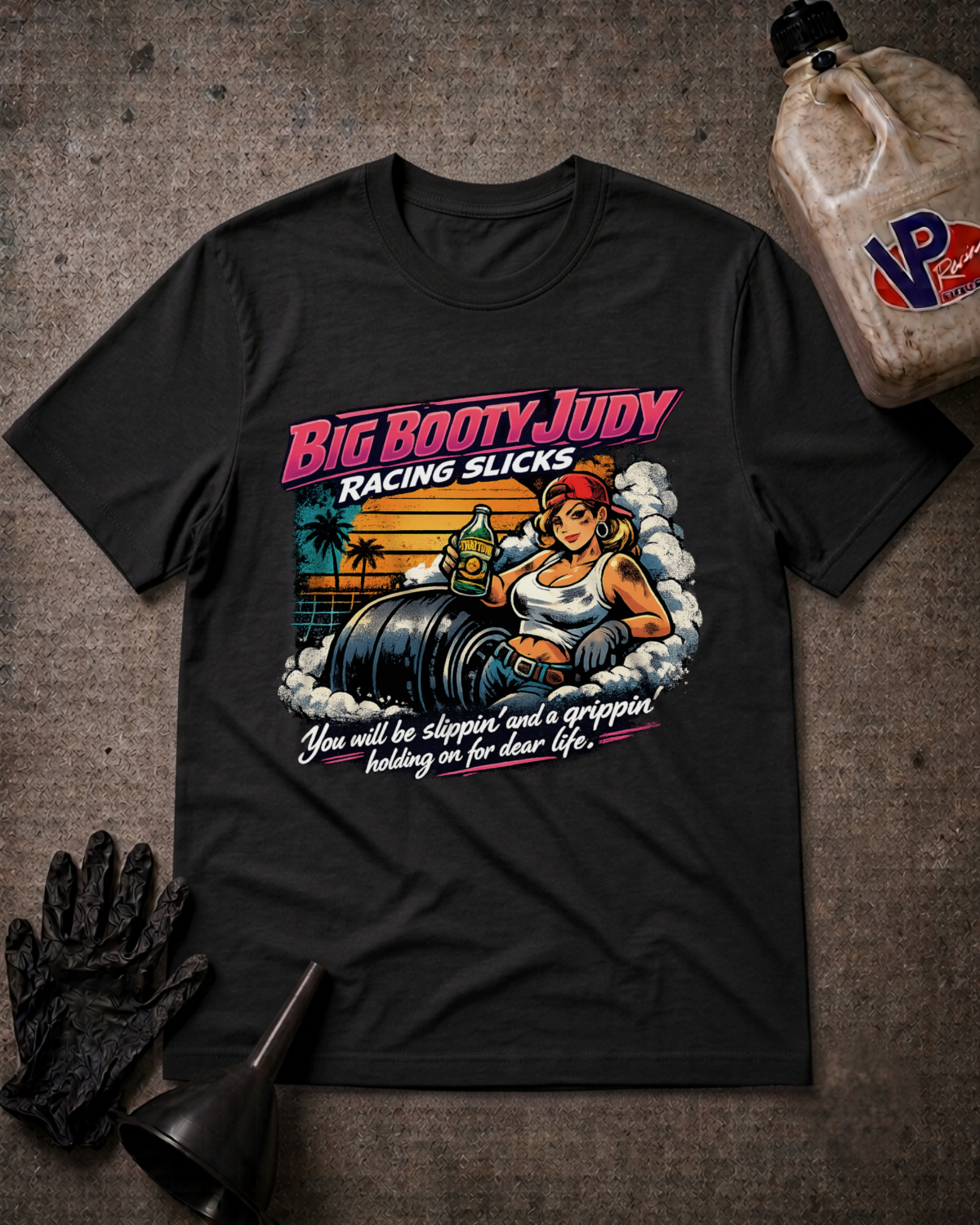 A black graphic t-shirt laid flat on a textured gray background, styled with a black glove, a funnel, and a plastic jug. The t-shirt features a colorful retro-style graphic of a woman labeled 'Big Booty Judy' sitting on race slicks with the text 'Racing Slicks' and 'You will be slippin' and a grippin holding on for dear life'.