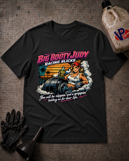 A black graphic t-shirt laid flat on a textured gray background, styled with a black glove, a funnel, and a plastic jug. The t-shirt features a colorful retro-style graphic of a woman labeled 'Big Booty Judy' sitting on race slicks with the text 'Racing Slicks' and 'You will be slippin' and a grippin holding on for dear life'.