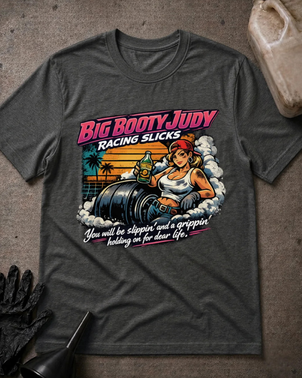 A dark heather gray graphic t-shirt laid flat on a textured gray surface, styled with a black work glove, a funnel, and a plastic jug. The t-shirt features a retro-style pin-up graphic of a woman in a racing slick sitting on a barrel, with the text 'BIG BOOTY JUDY RACING SLICKS' and 'You will be slipping' and 'a gripper' leading to 'dear life.