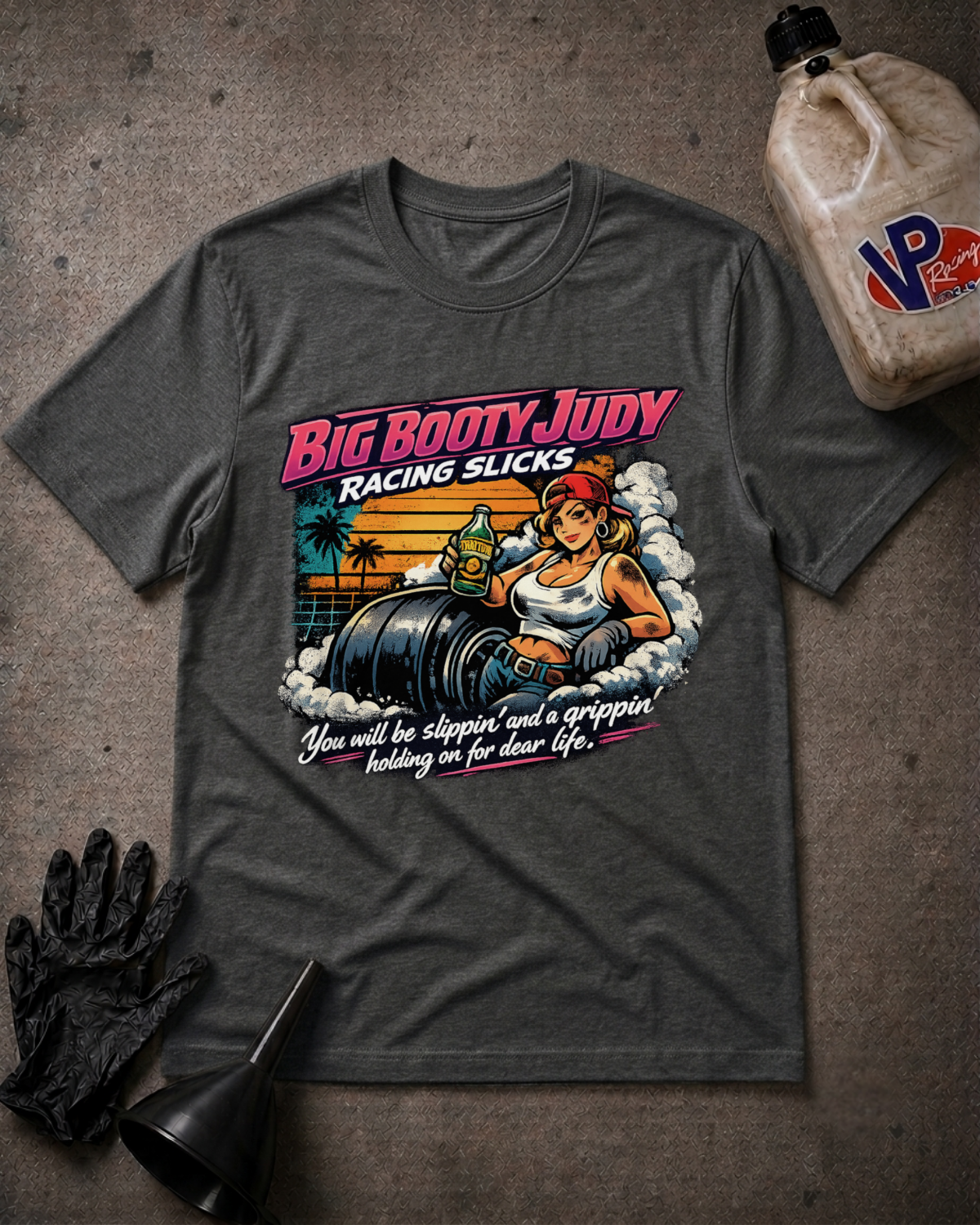 A dark gray graphic t-shirt laid flat on a textured gray surface, surrounded by themed props including a black racing glove, a funnel, and a plastic jug. The t-shirt features a retro-style graphic of a woman in a racing suit next to a race slicks with the text 'Big Booty Judy Racing Slicks'.