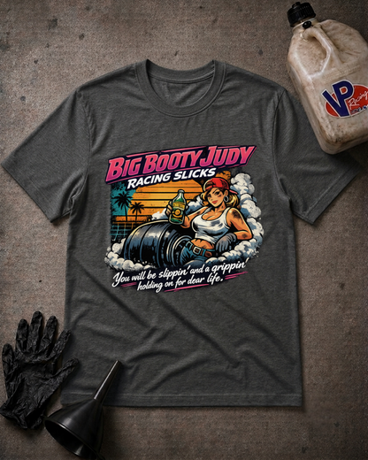 A dark gray graphic t-shirt laid flat on a textured gray surface, surrounded by themed props including a black racing glove, a funnel, and a plastic jug. The t-shirt features a retro-style graphic of a woman in a racing suit next to a race slicks with the text 'Big Booty Judy Racing Slicks'.
