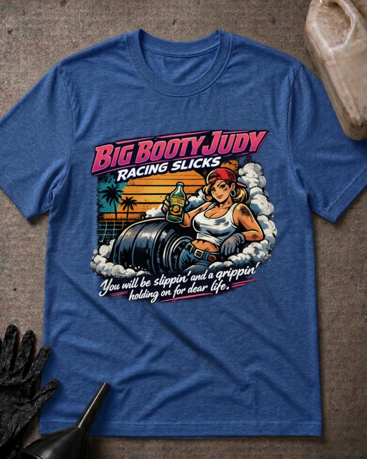 A heather blue graphic t-shirt laid flat on a textured gray surface. The shirt features a retro-style illustration of a woman in a red cap and white tank top next to a barrel, with the text 'BIG BOOTY JUDY RACING SLICKS' and 'You will be slippin' and a grippin' holding on for dear life.