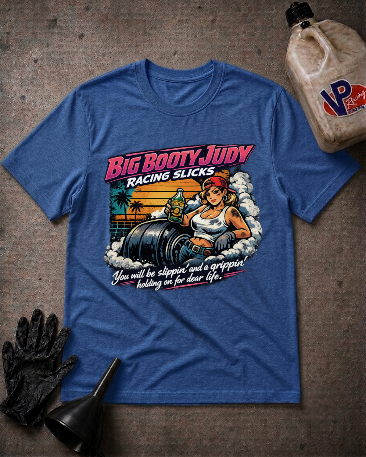 A heather blue graphic t-shirt laid flat on a textured gray surface, surrounded by racing-themed props including a black glove, funnel, and a jug of VP Racing. The shirt features a retro-style graphic of a woman in a racing suit with the text 'BIG BOOTY JUDY RACING SLICKS' and 'You will be slippin' and a grippin holding on for dear life.