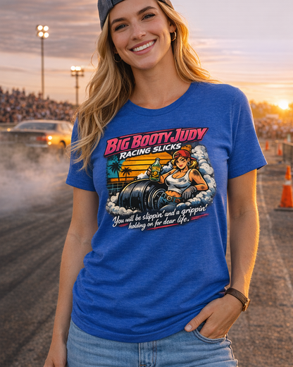 Woman wearing a blue t-shirt with a graphic design and 'Big Booty Judy Racing Slicks You will be slippin' and a grippin' holding on for dear life' text at a race track.
