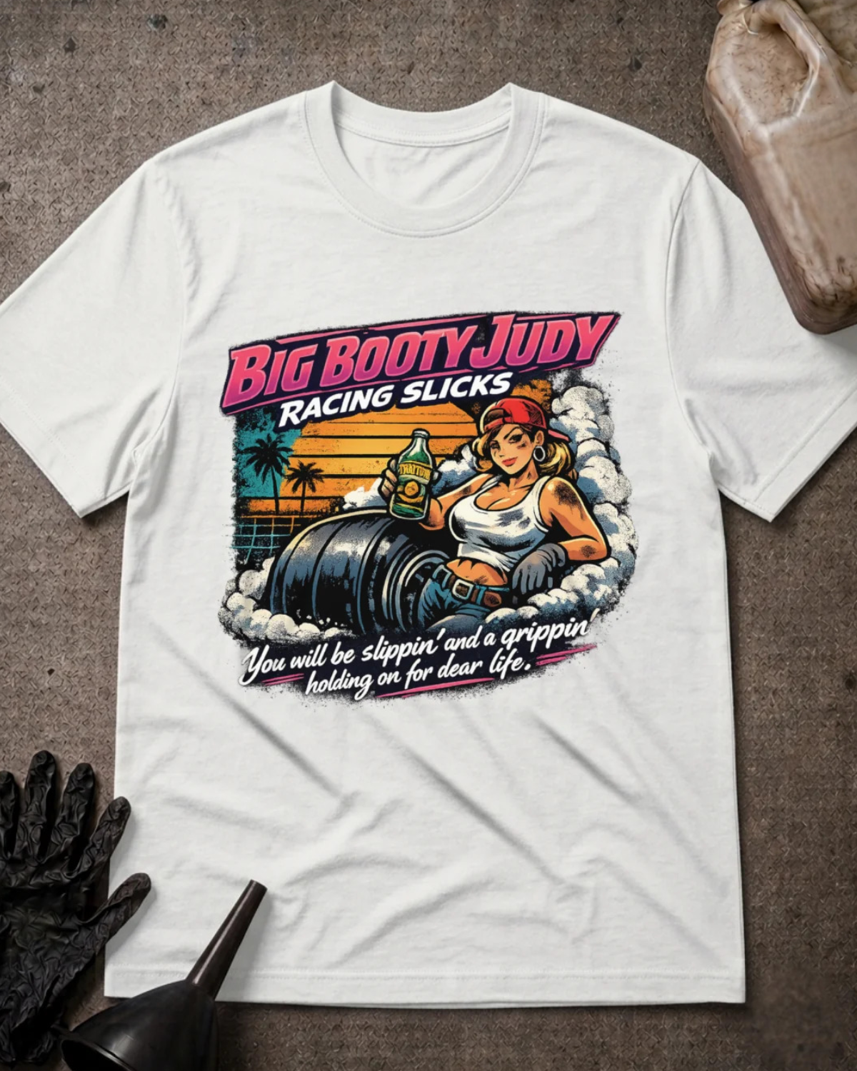 A white graphic t-shirt laid flat on a textured gray surface, styled with a black work glove, a funnel, and a plastic jug. The t-shirt features a retro-style pin-up graphic of a woman in a racing slick sitting on a barrel, with the text 'BIG BOOTY JUDY RACING SLICKS' and 'You will be slipping' and 'a gripper' leading to 'dear life.