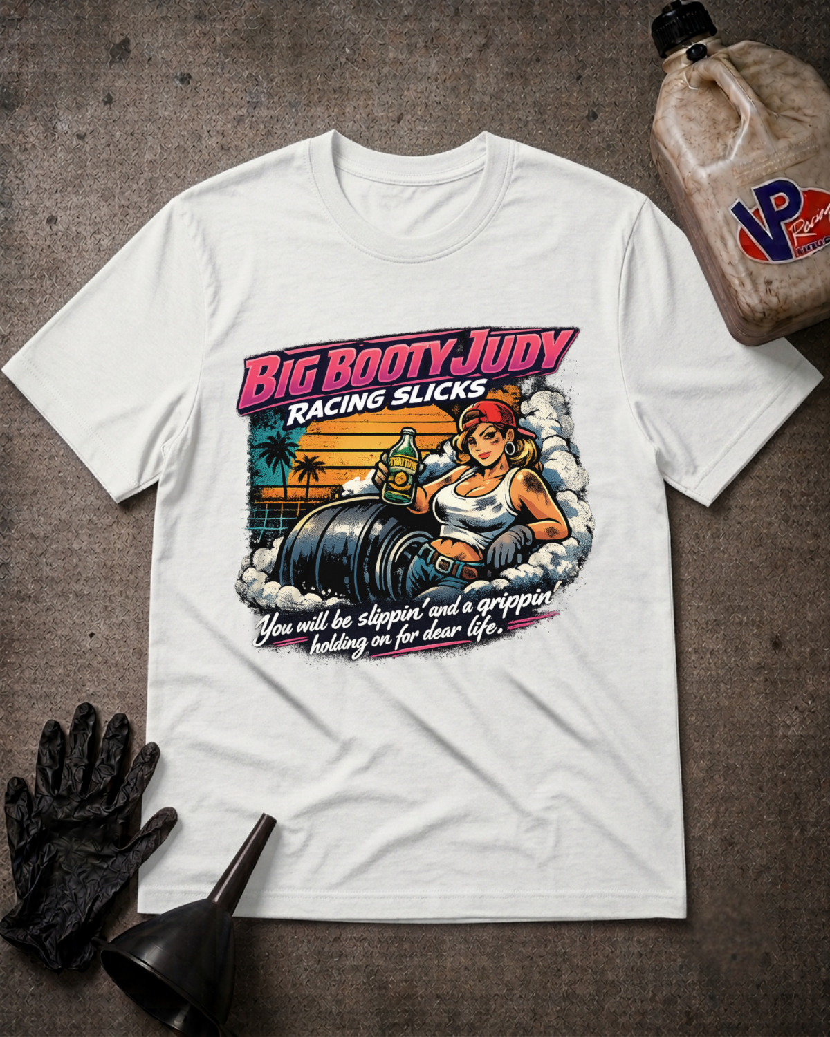 A white graphic t-shirt featuring a retro-style illustration of a woman in a racing suit with the text 'Big Booty Judy Racing Slicks' and 'You will be slippin' and a gripping holding on for dear life'. The shirt is displayed in a flat-lay arrangement on a textured gray background, accompanied by a black glove, a black funnel, and a bottle of VP Racing Slicks.