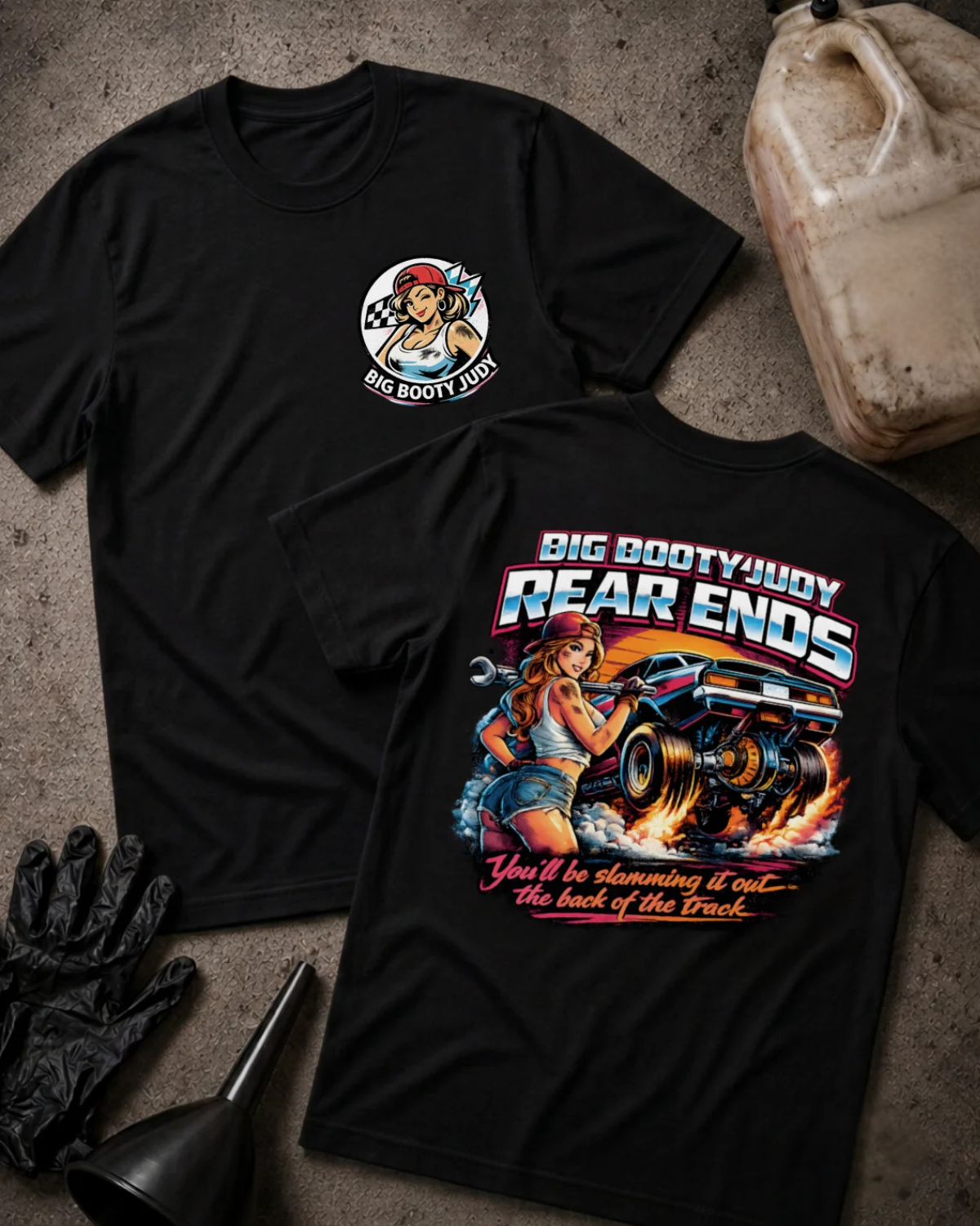 Two black graphic T-shirts laid flat on a textured gray surface, with automotive-themed artwork and text. One shirt shows a circular chest graphic, and the other shows a large back graphic with the words “BIG BOOTY JUDY REAR ENDS” and “You’ll be slamming it out~ the back of the track.