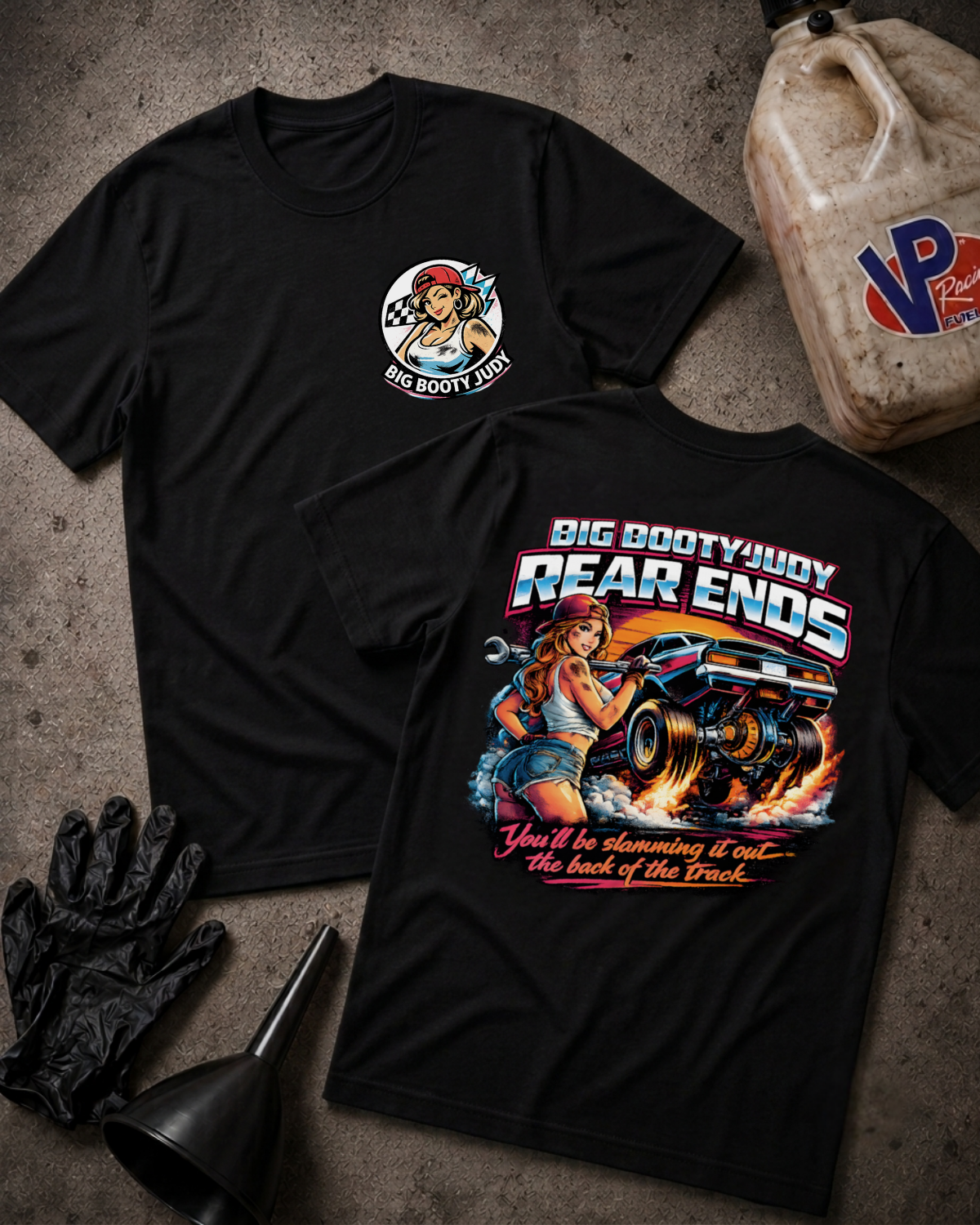Two black graphic T-shirts laid flat on a textured gray surface, with automotive-themed artwork and text. One shirt shows a circular chest graphic, and the other shows a large back graphic with the words “BIG BOOTY JUDY REAR ENDS” and “You’ll be slamming it out~ the back of the track.