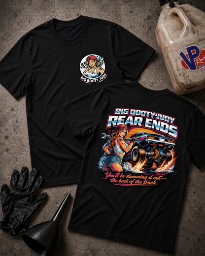 Two black graphic T-shirts laid flat on a textured gray surface, with automotive-themed artwork and text. One shirt shows a circular chest graphic, and the other shows a large back graphic with the words “BIG BOOTY JUDY REAR ENDS” and “You’ll be slamming it out~ the back of the track.