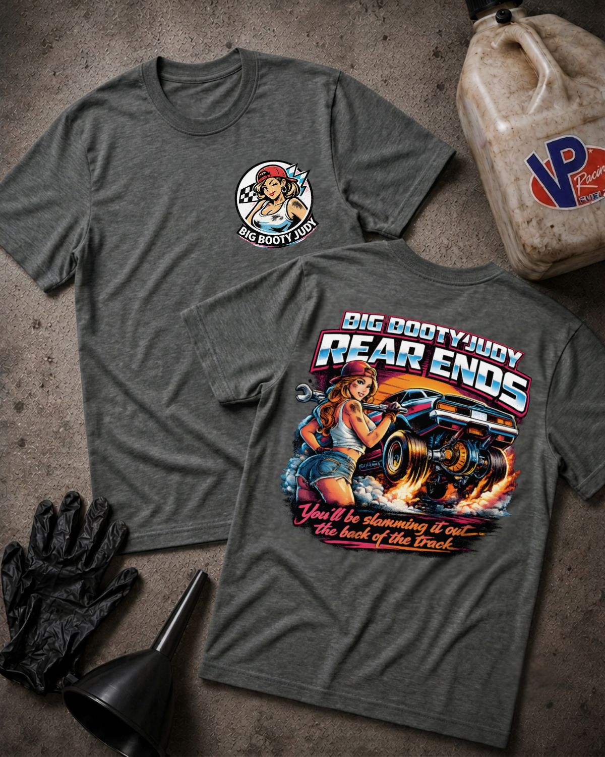 Two heather dark gray graphic T-shirts laid flat on a textured gray surface, showing front and back designs. The front has a small circular 'Big Booty Judy' logo on the chest, and the back has a large colorful graphic with the text 'Big Booty Judy Rear Ends' and 'You'll be slamming it out the back of the Track.