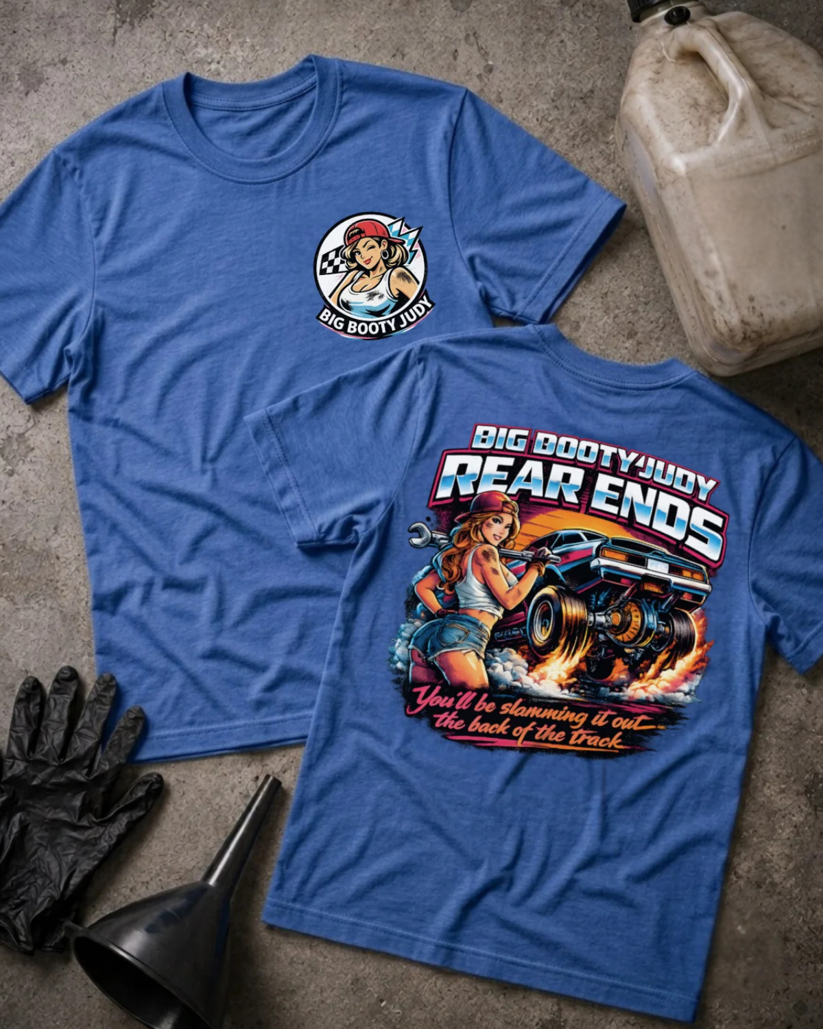 Two blue graphic T-shirts laid flat on a concrete surface, with automotive-themed artwork and text. One shirt shows a small circular chest graphic, and the other shows a large back graphic reading 'Big Booty Judy Rear Ends' with a woman, car, and flames.