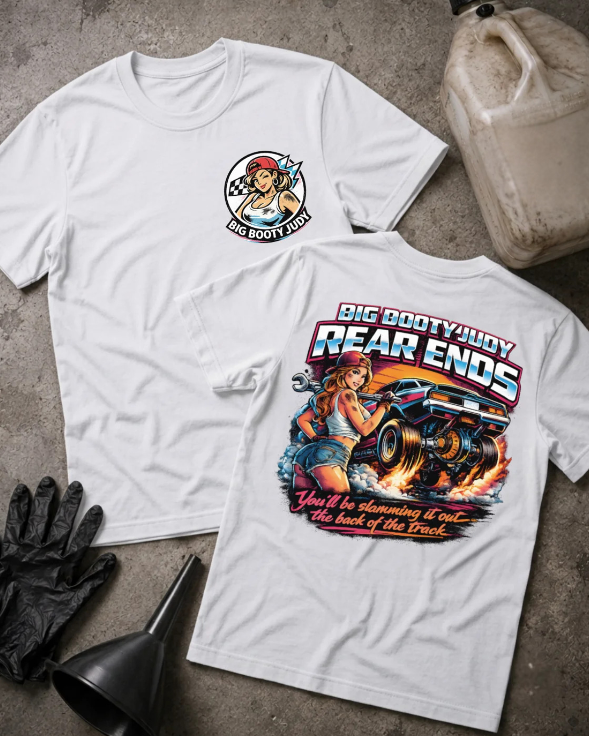 Two white aphic T-shirts laid flat on a textured gray surface, with automotive-themed artwork and text. One shirt shows a circular chest graphic, and the other shows a large back graphic with the words “BIG BOOTY JUDY REAR ENDS” and “You’ll be slamming it out~ the back of the track.