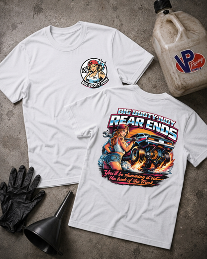 Two white graphic T-shirts laid flat on a gray concrete surface, with a black funnel, black gloves, and a plastic jug nearby. The front shirt has a small circular chest graphic, and the back shirt has a large colorful graphic reading 'Big Booty Judy Rear Ends' with an illustrated woman and car.