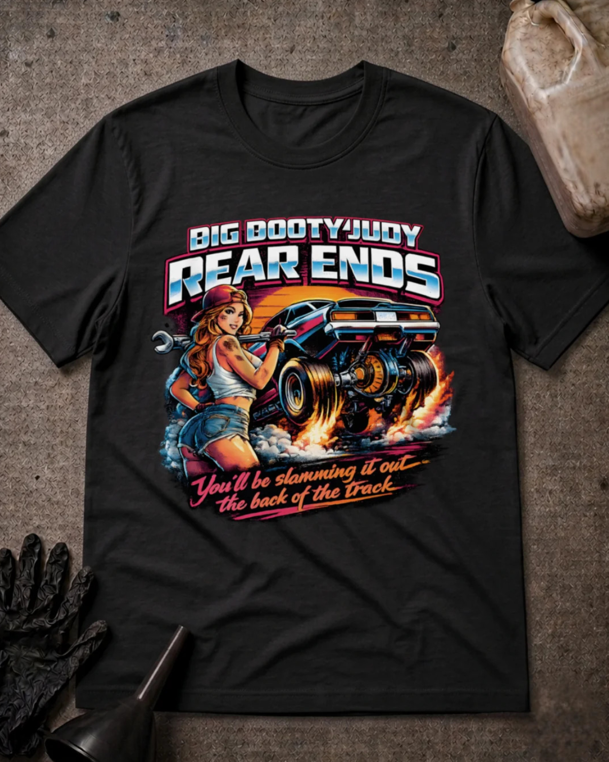 A black graphic t-shirt laid flat on a textured gray surface with automotive props. The graphic features a pin-up style illustration of a woman holding a wrench next to a race car, with the text 'BIG BOOTYJUDY REAR ENDS' and 'You'll be slamming it out the back of the Track'.