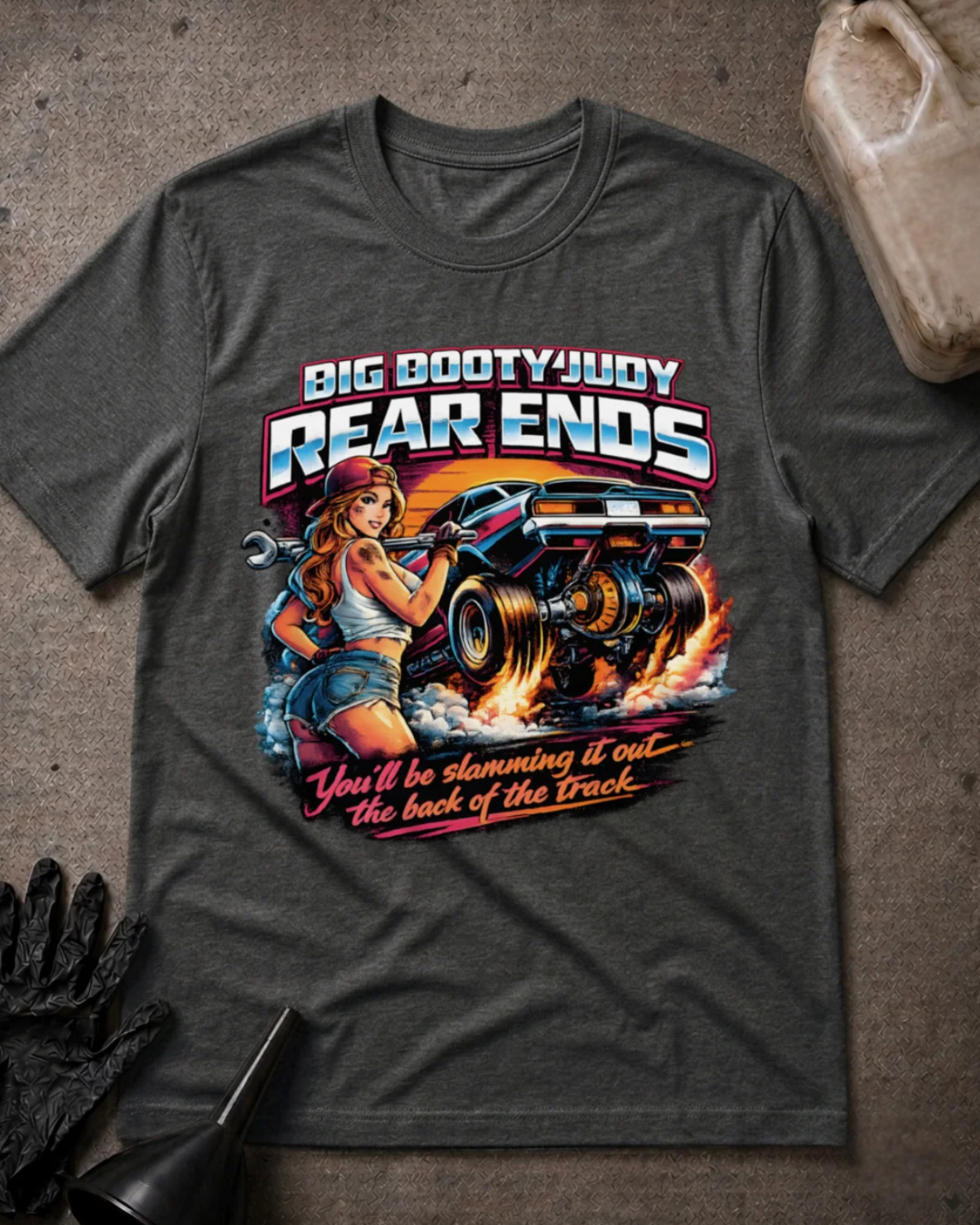 A dark grey heather  graphic t-shirt laid flat on a textured gray surface with automotive props. The graphic features a pin-up style illustration of a woman holding a wrench next to a race car, with the text 'BIG BOOTYJUDY REAR ENDS' and 'You'll be slamming it out the back of the Track'.